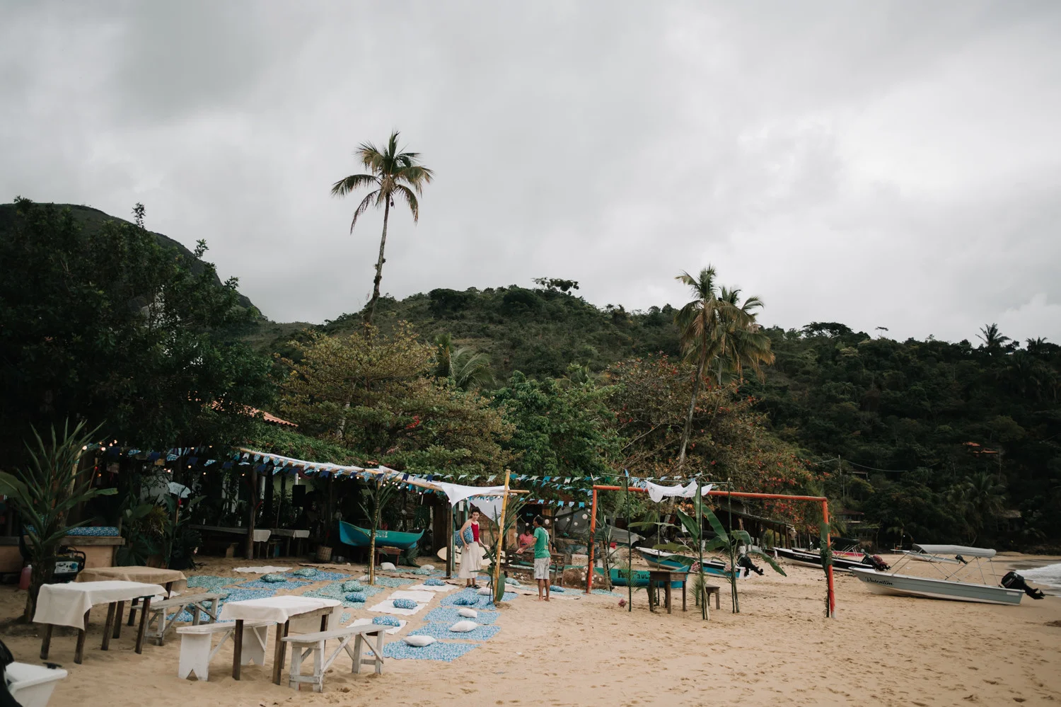 Beach wedding setup at Pouso das Flores in Paraty, Brazil, with colorful bunting, pillows and white tables on sand.