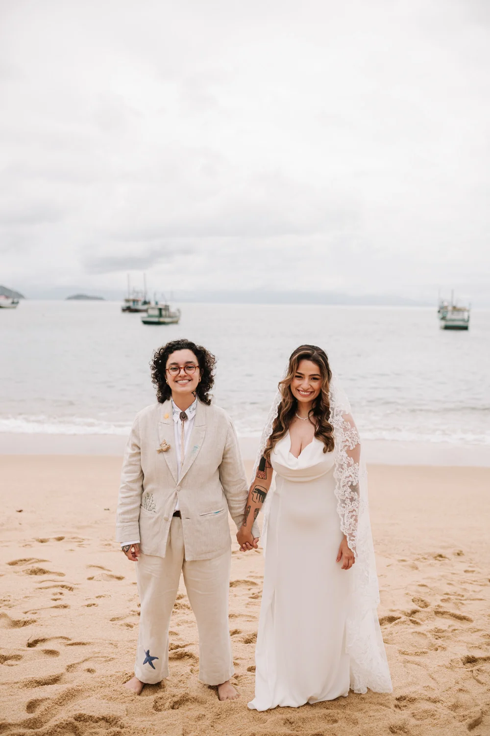 Rafaela and Nathalia hold hands on the beach at Pouso das Flores in Paraty, Brazil, smiling in wedding attire.