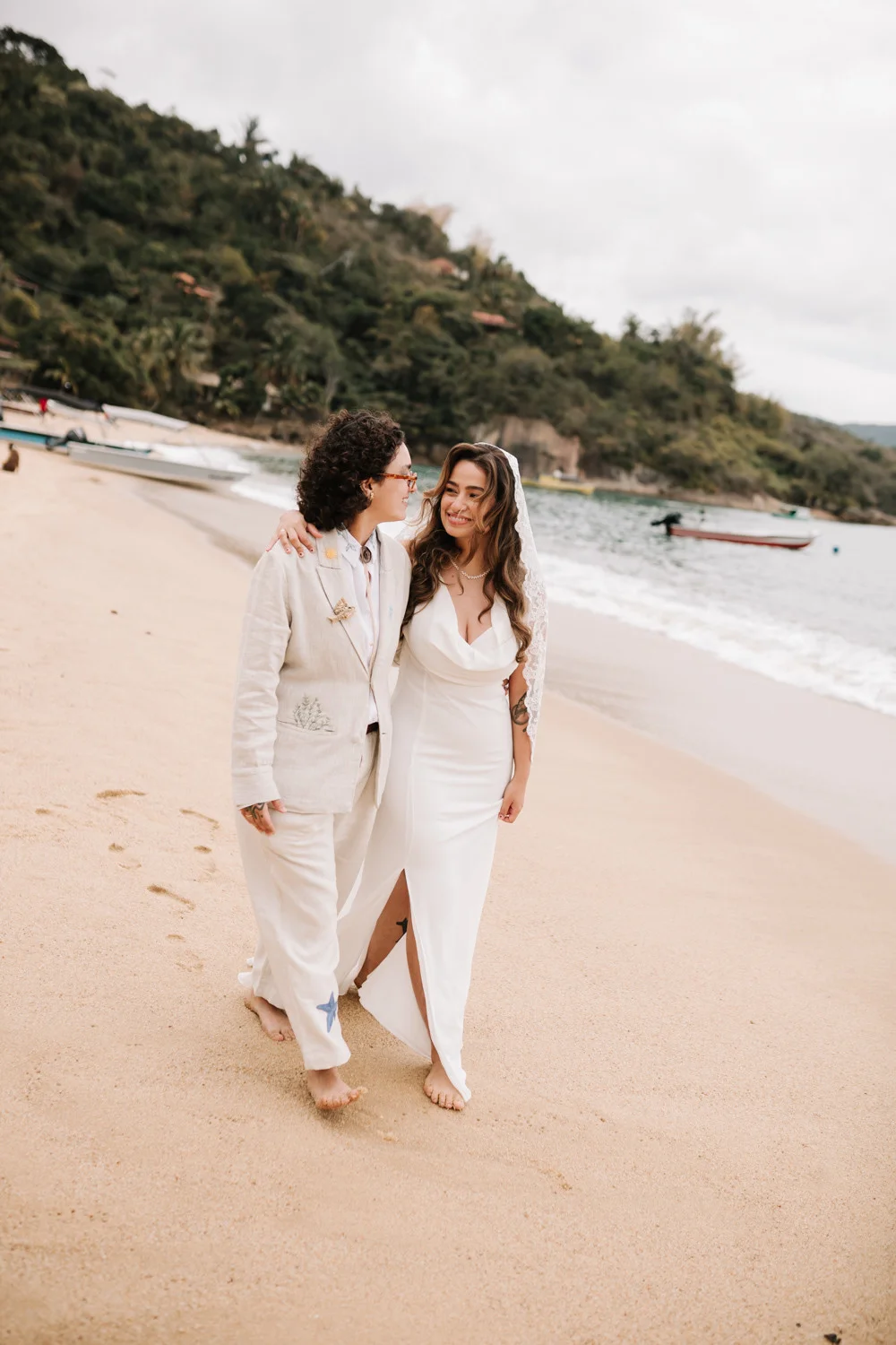 Rafaela and Nathalia walk barefoot along a sandy beach in Paraty, Brazil, smiling at each other on their wedding day.