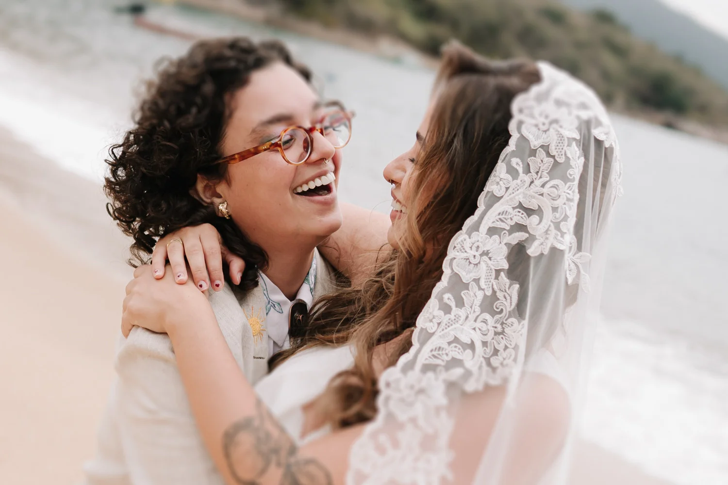 Rafaela and Nathalia laugh joyfully on the beach at Pouso das Flores in Paraty, Brazil, one wearing a lace veil.