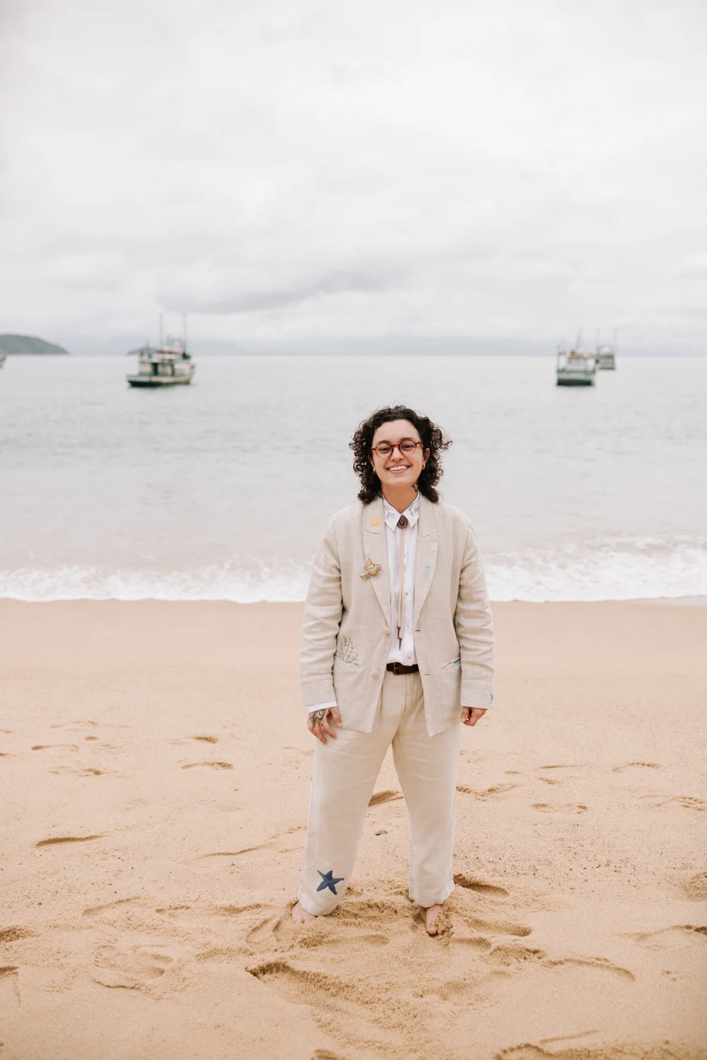 Rafaela smiles on the beach at Pouso das Flores in Paraty, Brazil, wearing a linen suit with starfish embroidery.