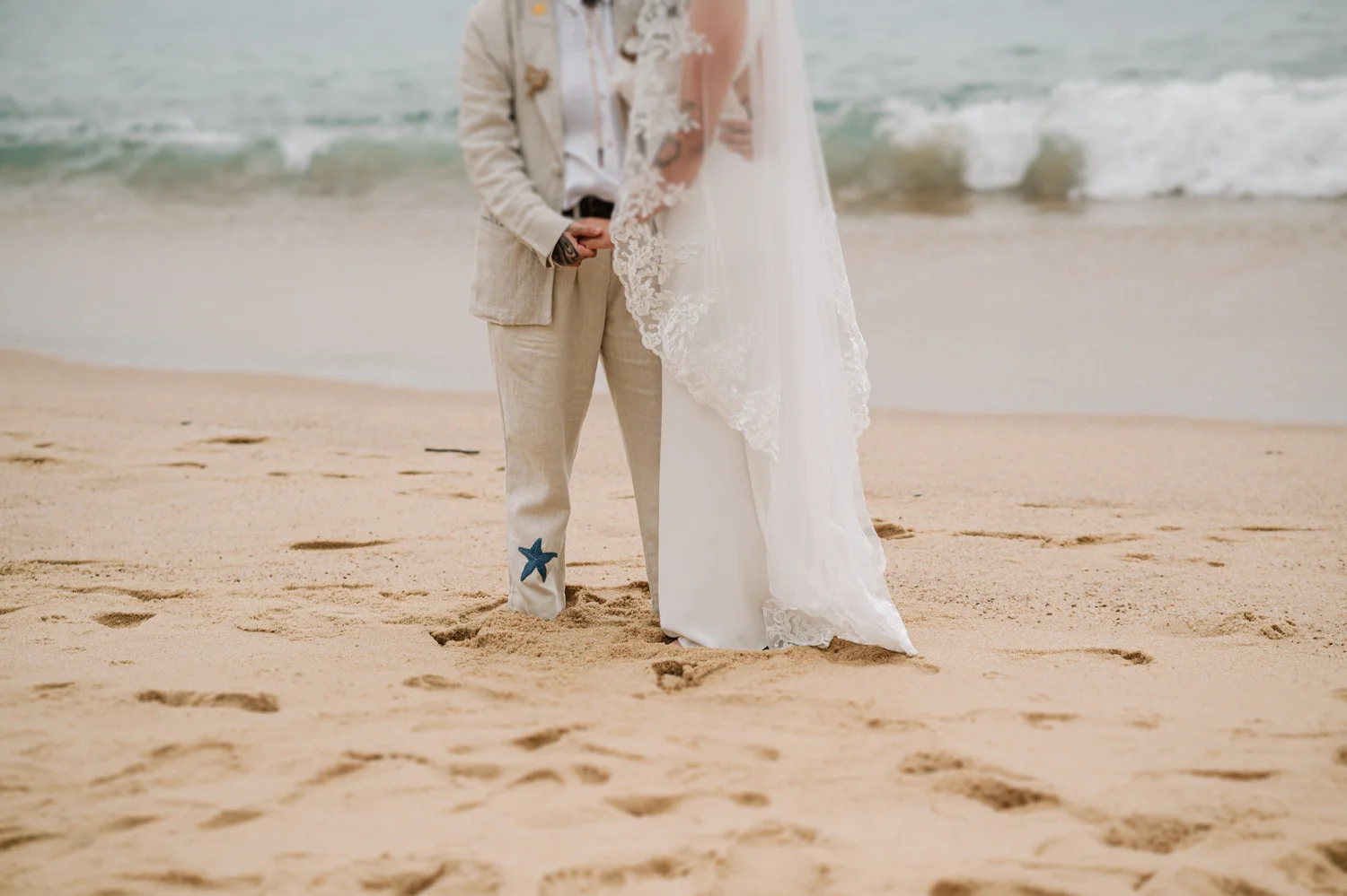 Rafaela and Nathalia hold hands on the beach at Pouso das Flores in Paraty, Brazil, barefoot in the sand.