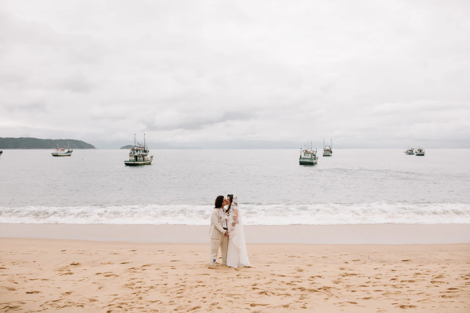 Rafaela and Nathalia share a kiss on the beach at Pouso das Flores in Paraty, Brazil, with fishing boats anchored offshore.