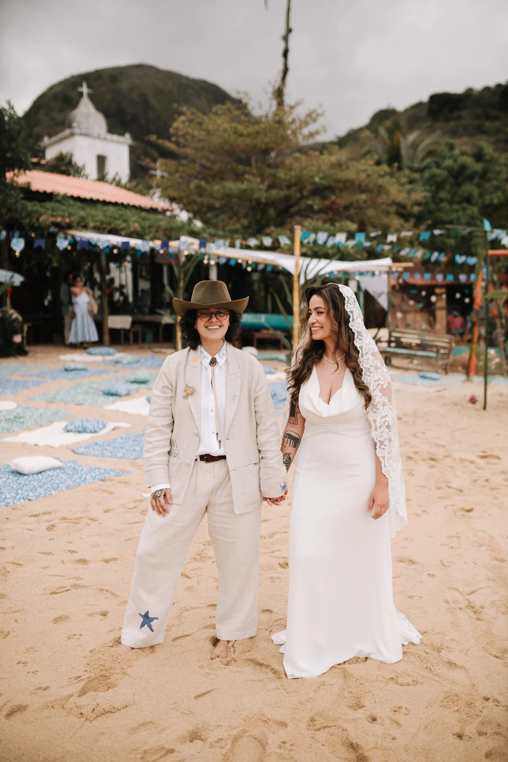 Rafaela and Nathalia hold hands smiling on sandy beach at Pouso das Flores in Paraty, Brazil.