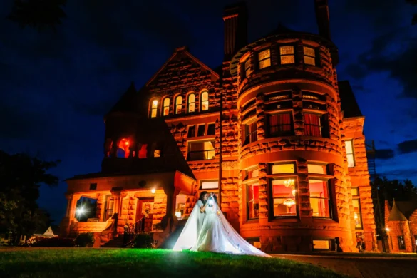 Bride Vanessa Crain holds a glowing lantern in a white gown before the illuminated Orman Mansion at night in Pueblo, Colorado.