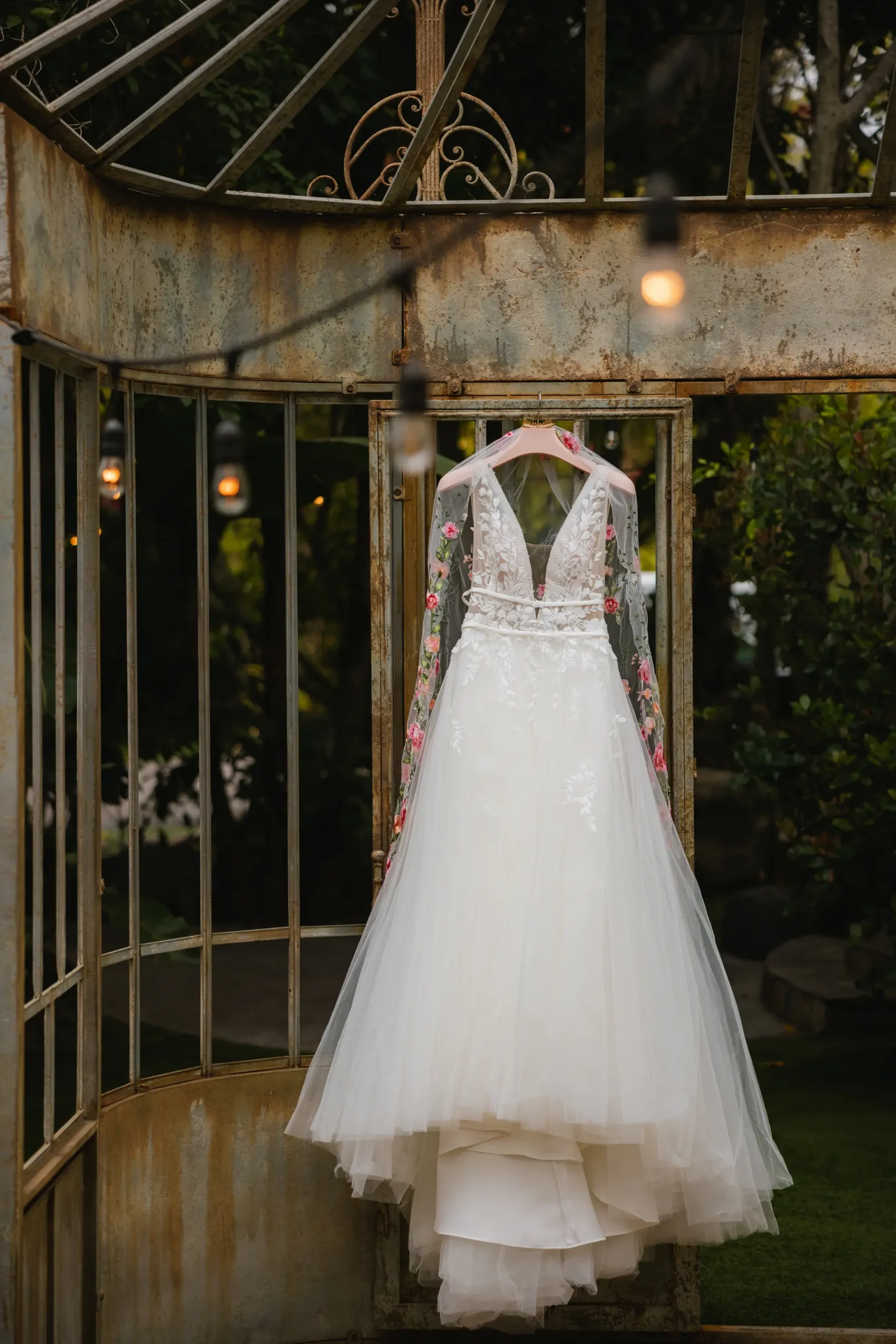 Floral-embroidered wedding gown with tulle skirt hangs from a pink hanger inside a rustic iron greenhouse at Botanica in Oceanside, California.