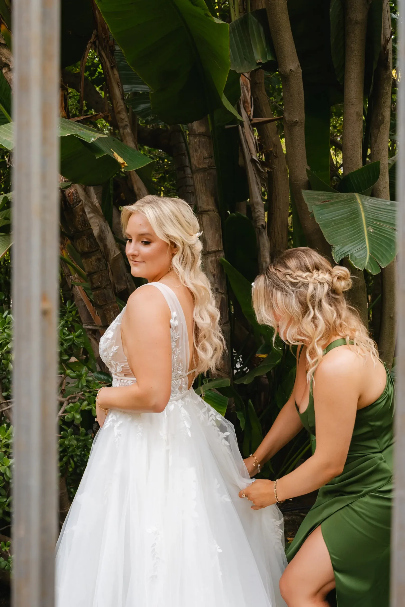 Bridesmaid in green dress fastens the back of bride Erika Beursken's white lace gown amid tropical foliage at Botanica in Oceanside, California.