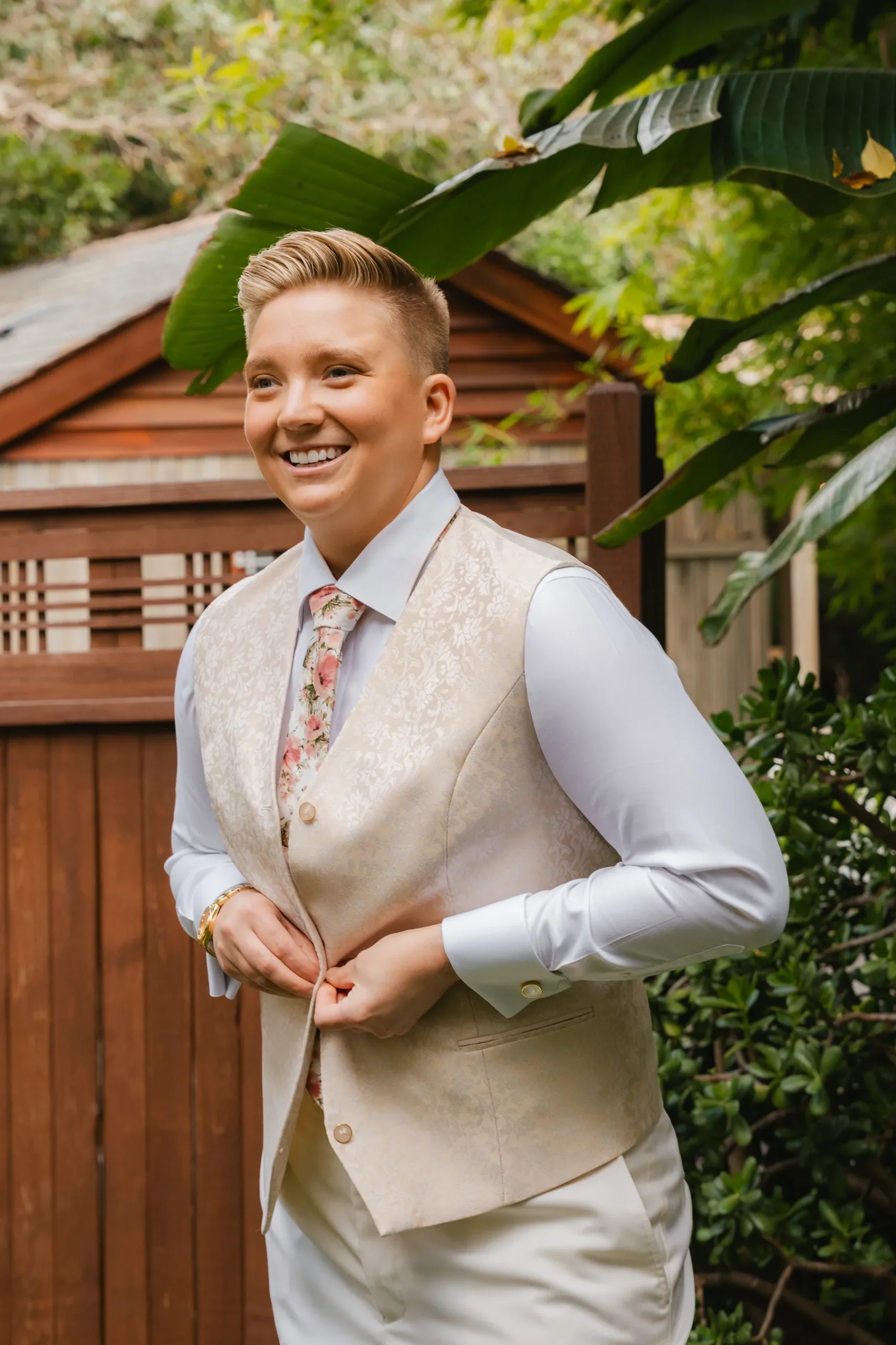 A smiling person in a cream brocade vest and floral tie buttons up while getting ready at Botanica in Oceanside, California.