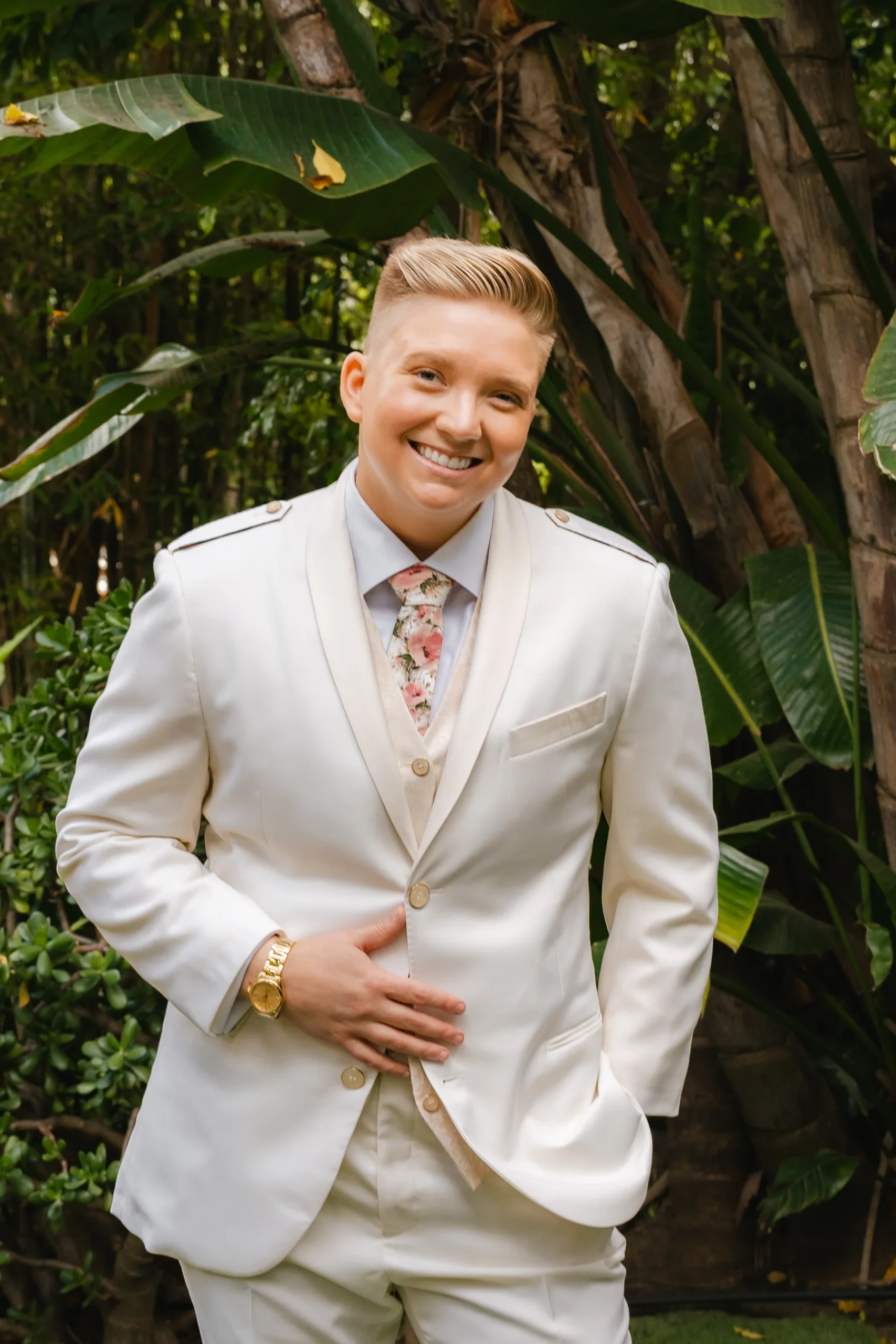 A smiling person in a white suit and floral tie poses outdoors at Botanica in Oceanside, California.