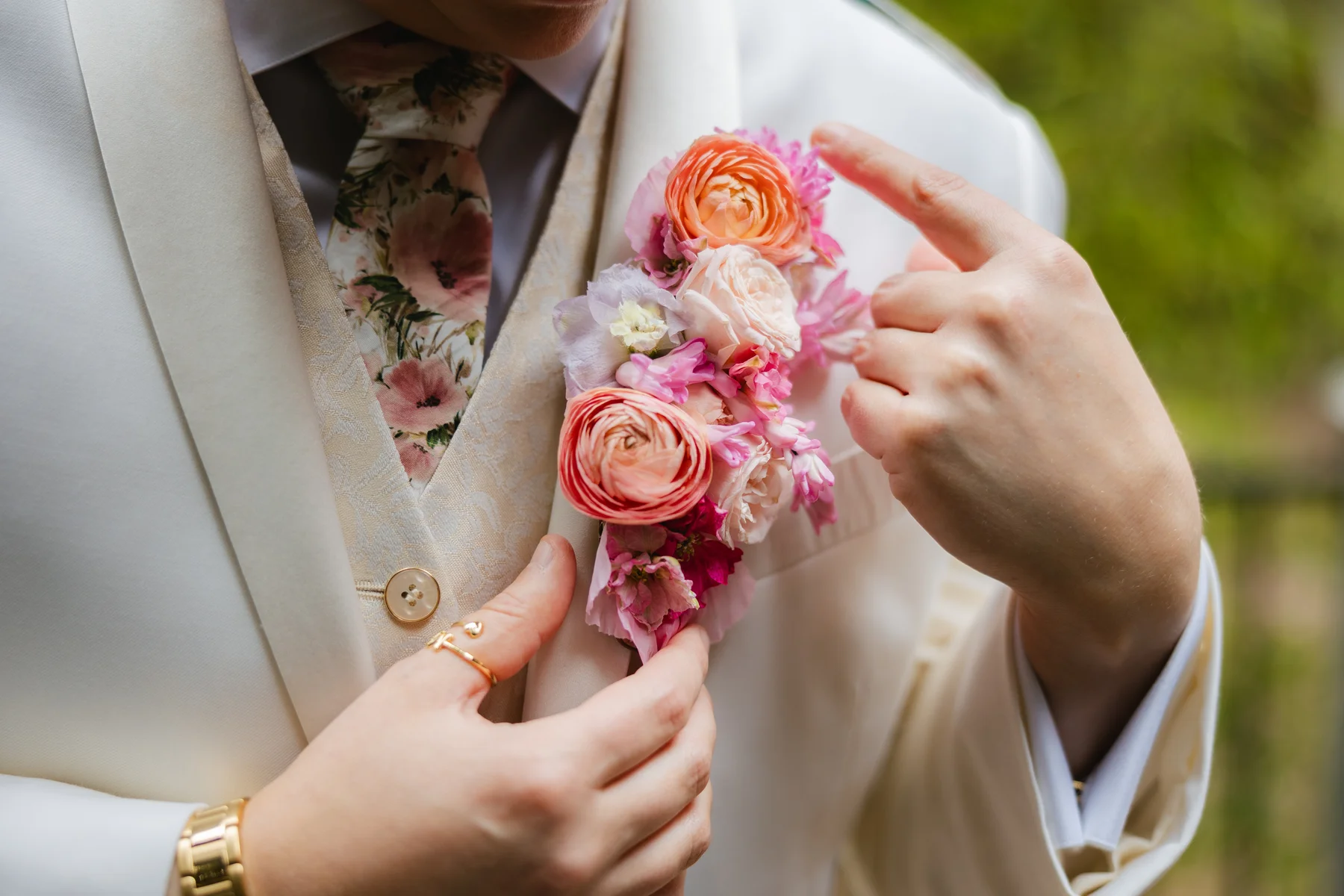 Hands pin a lush pink and coral ranunculus boutonniere on a white suit lapel at Botanica in Oceanside, California.