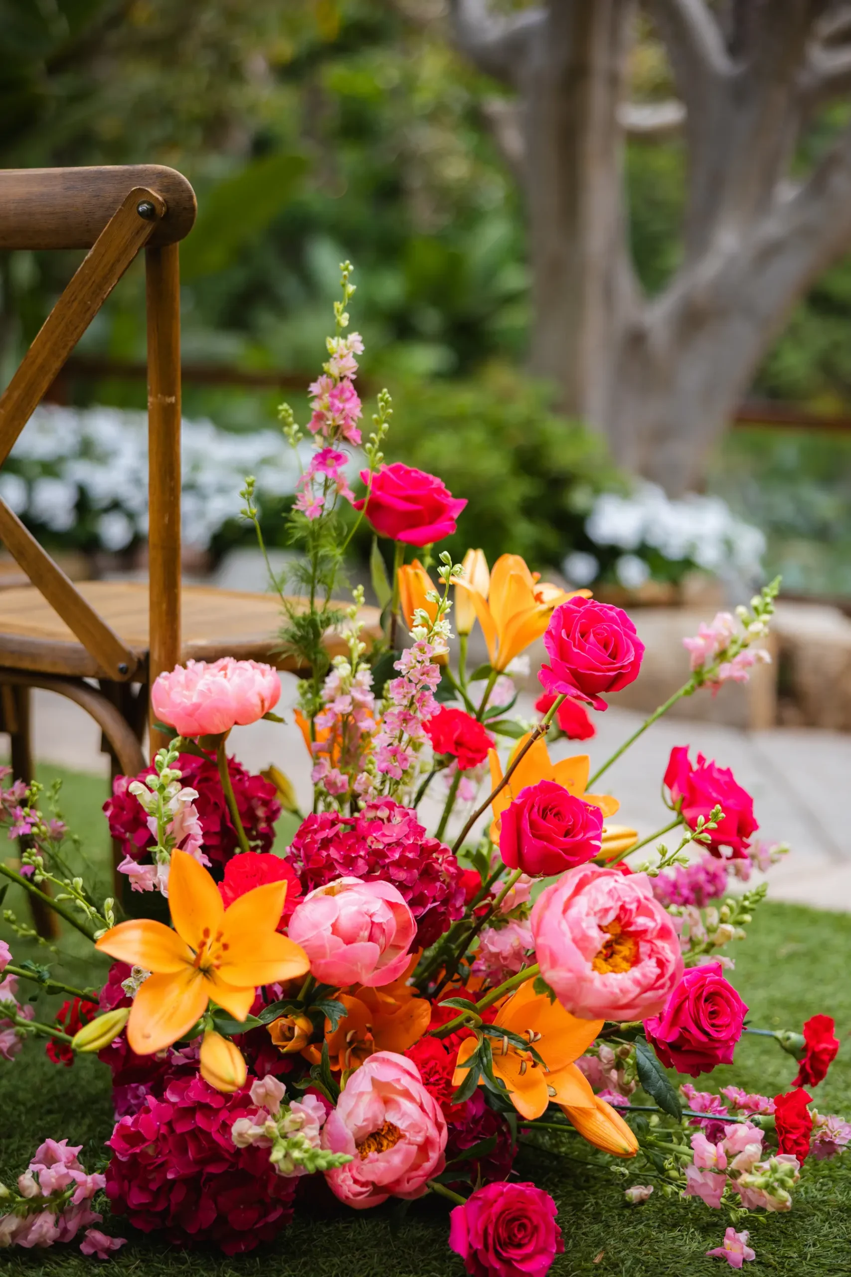 Lush floral arrangement of pink peonies, hot pink roses, orange lilies and magenta hydrangeas beside a wooden chair at Botanica in Oceanside, California.