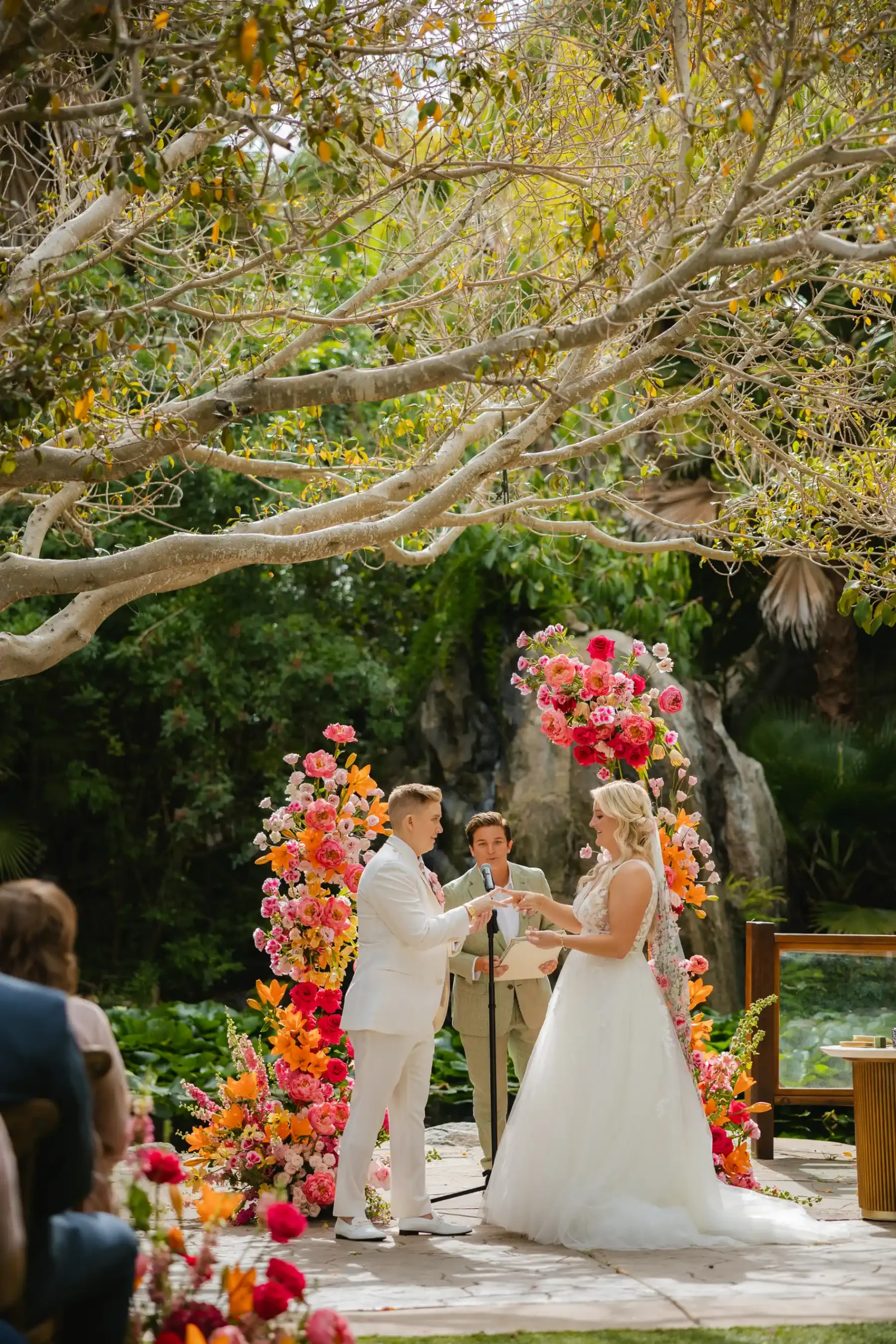 Erika Beursken and Melissa Brodsky exchange rings at Botanica in Oceanside, California, framed by a vibrant floral arch.