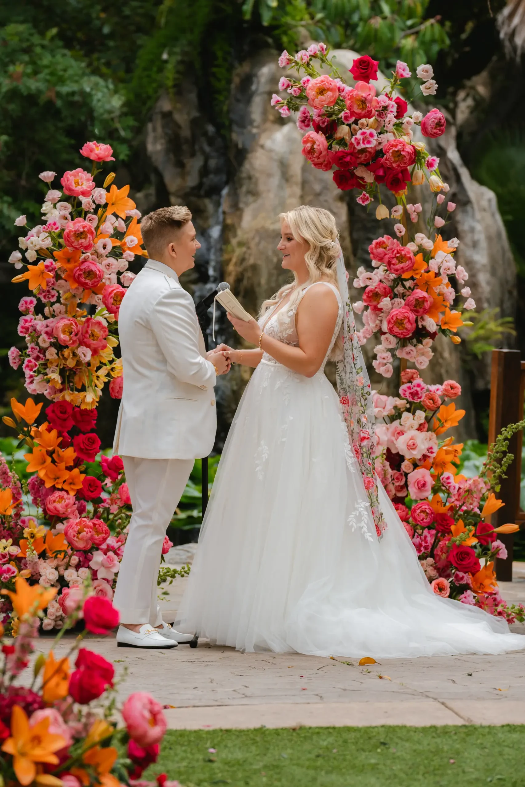 Erika Beursken and Melissa Brodsky exchange vows beneath a vibrant floral arch at Botanica in Oceanside, California.