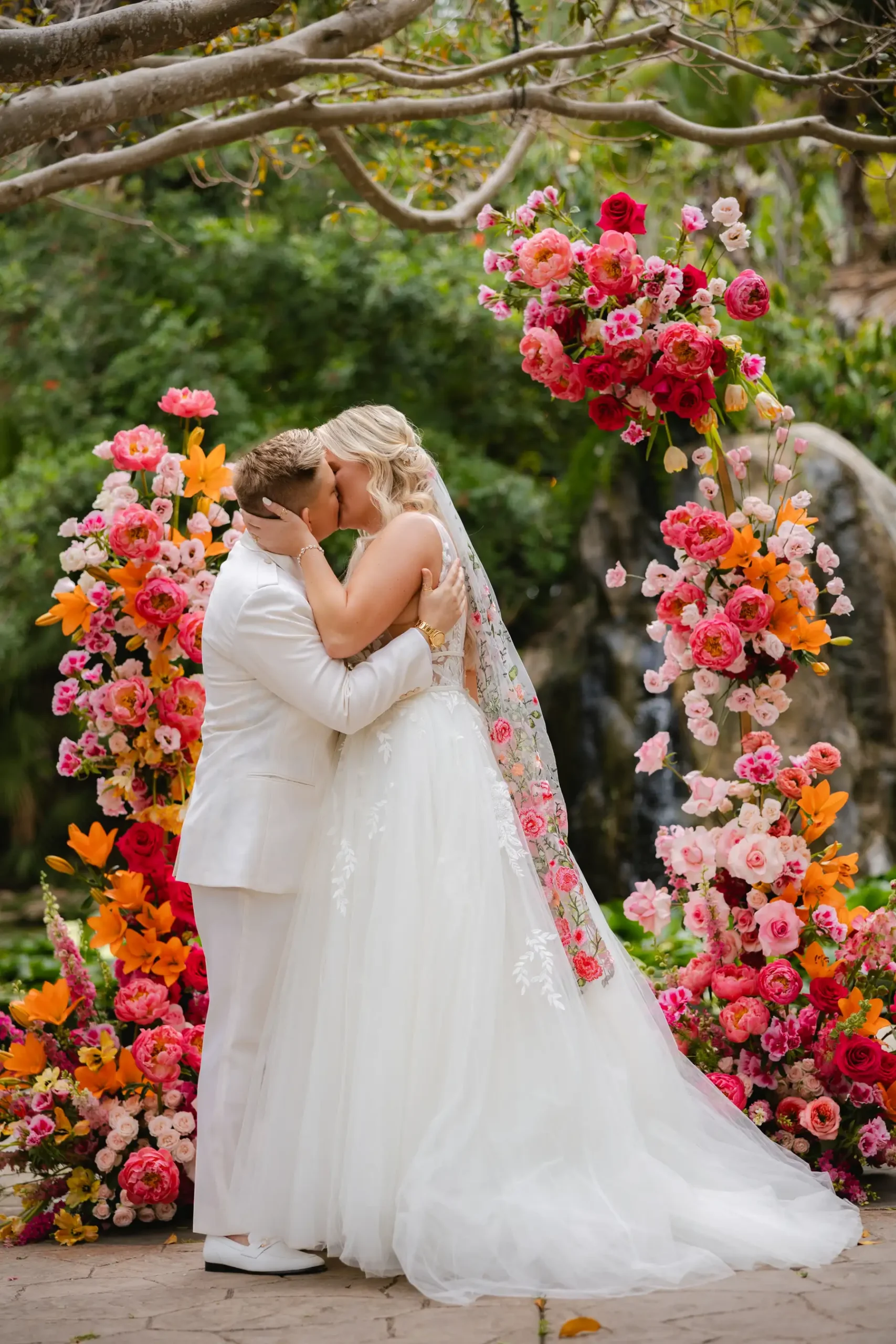Erika Beursken and Melissa Brodsky share their first kiss at Botanica in Oceanside, California, framed by a vibrant floral arch.