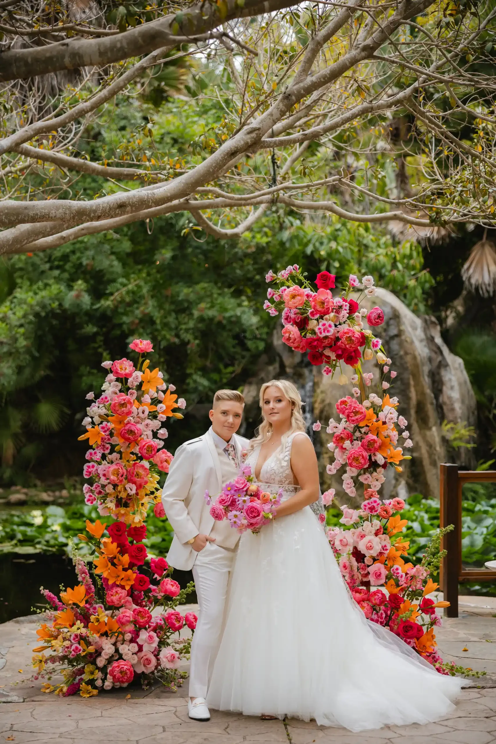 Erika Beursken and Melissa Brodsky pose before a vibrant floral arch at Botanica in Oceanside, California.