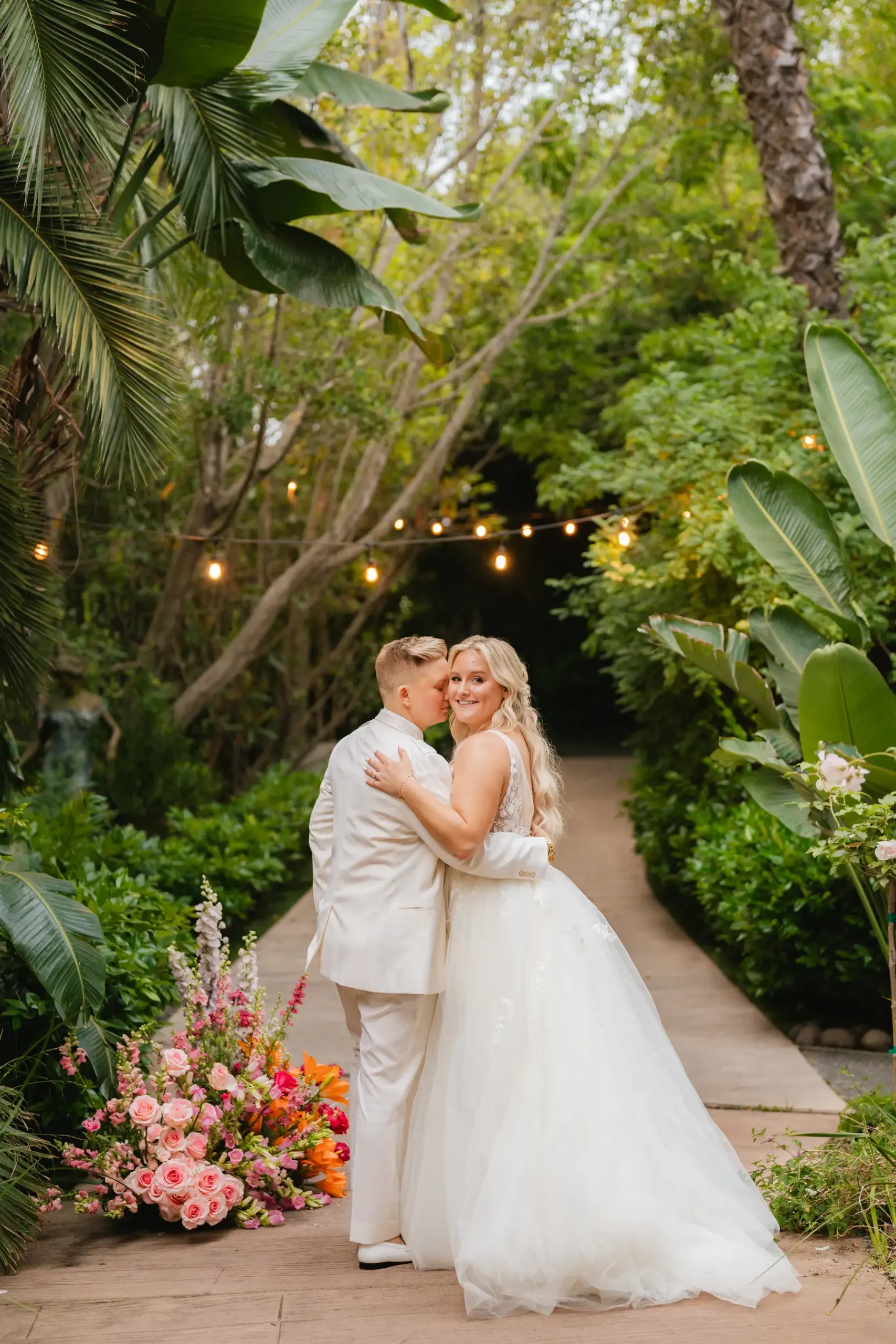 Erika Beursken and Melissa Brodsky embrace on a garden path at Botanica in Oceanside, California, surrounded by lush tropical greenery and colorful florals.