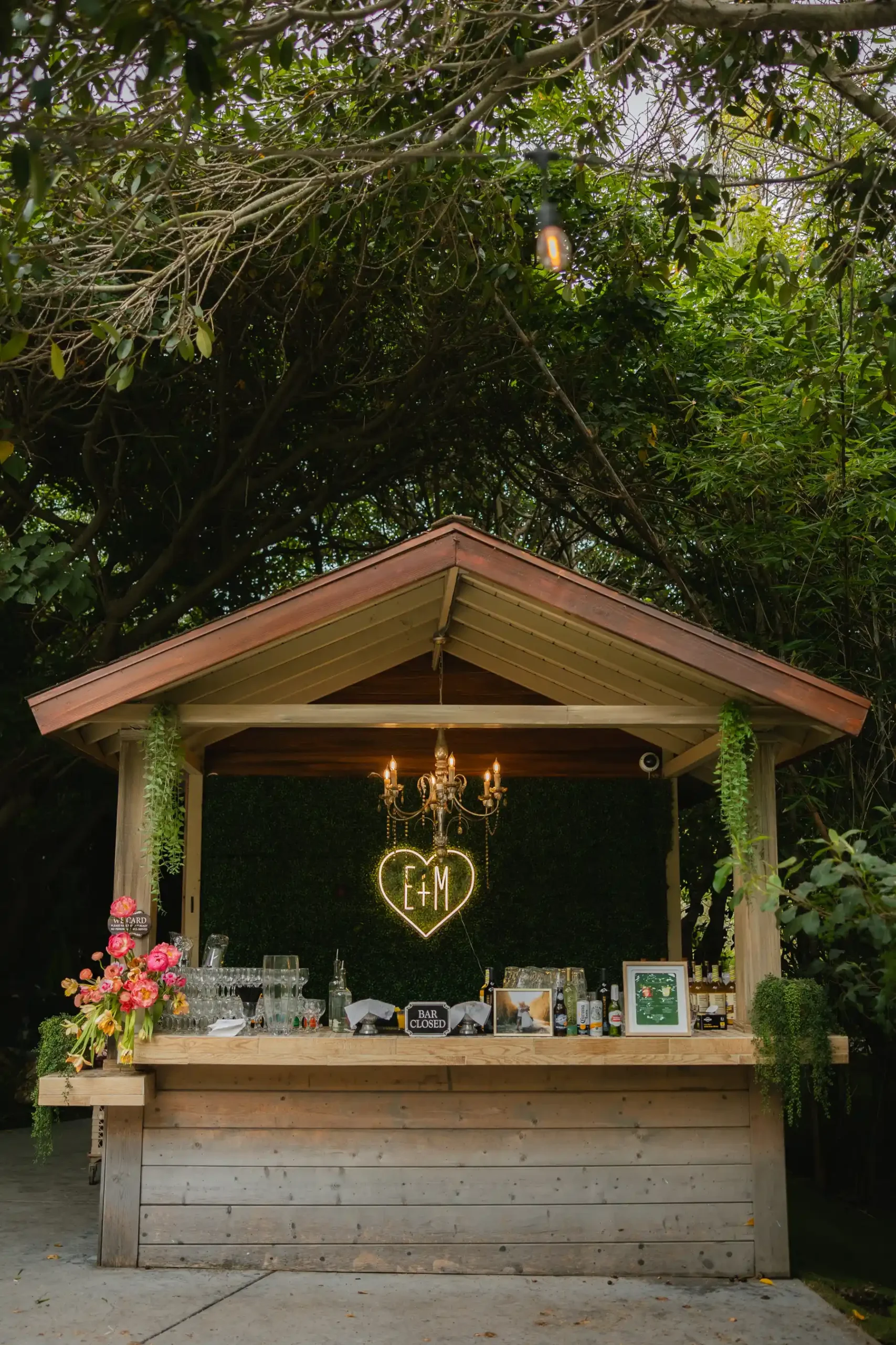 Outdoor wedding bar kiosk with neon E+M heart sign and chandelier at Botanica in Oceanside, California.