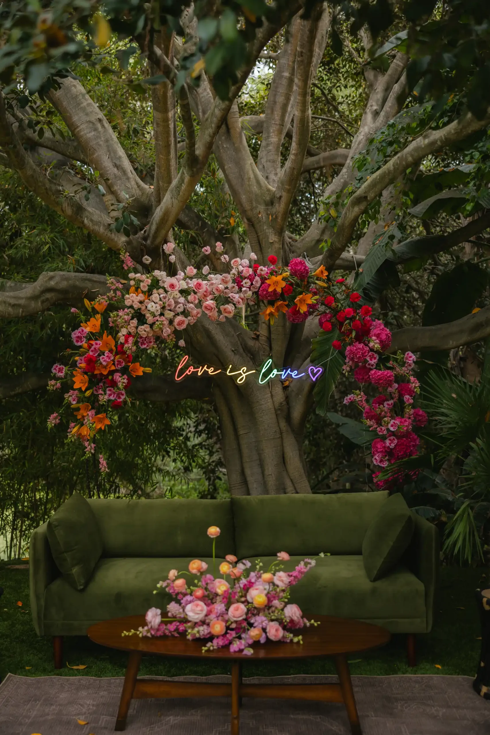 Floral arch with pink, red and orange blooms frames a 'love is love' neon sign above a green velvet sofa at Botanica in Oceanside, California.