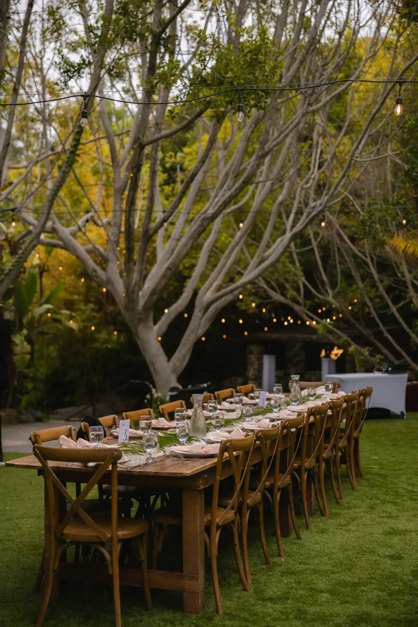 Long wooden farm table set for an outdoor wedding reception at Botanica in Oceanside, California, with string lights and lush greenery.