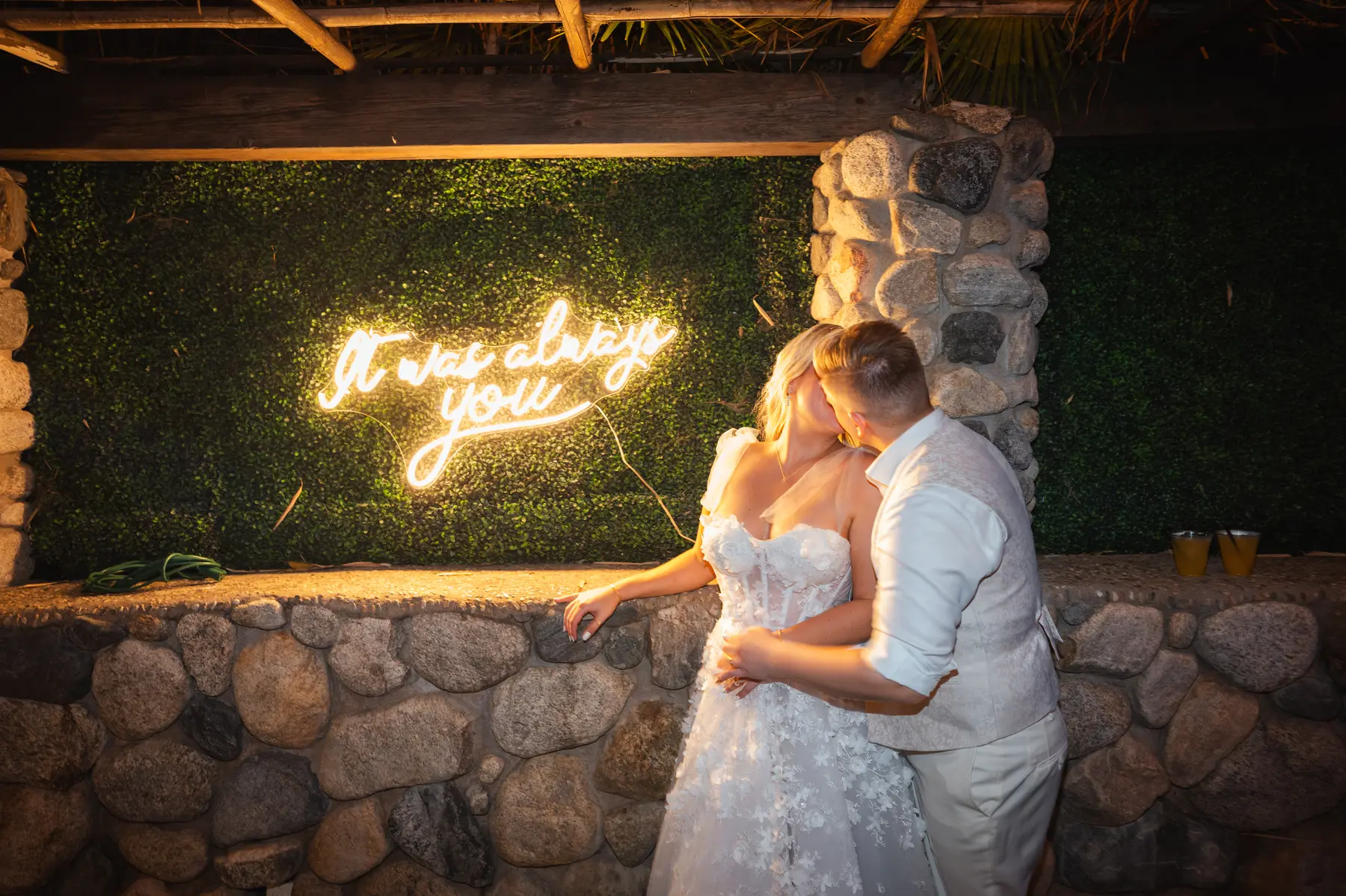 Erika Beursken and Melissa Brodsky share a kiss beside a glowing 'It was always you' neon sign at Botanica in Oceanside, California.