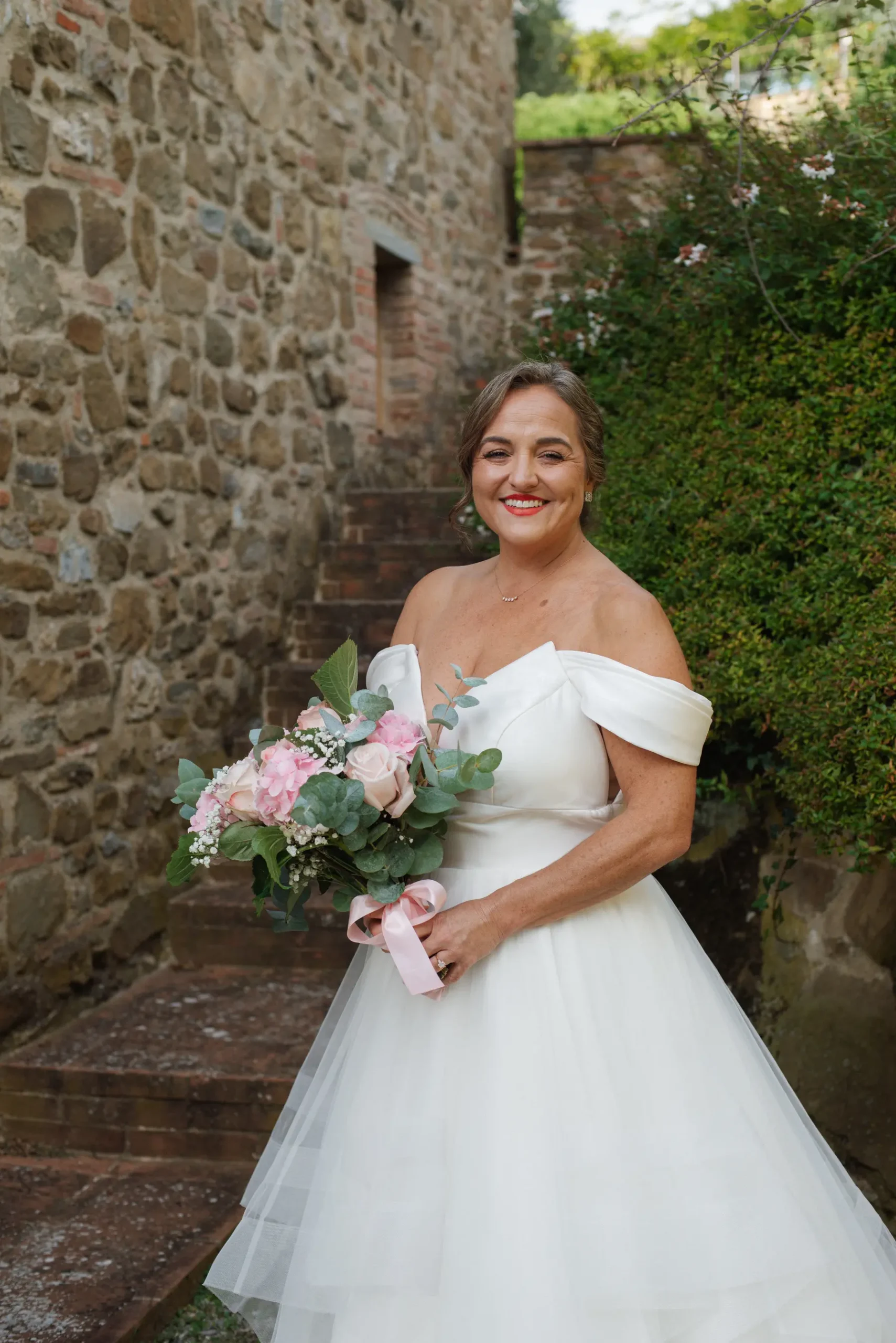 Bride Lauren Thomas smiles in an off-shoulder white gown holding a pink floral bouquet on stone steps at Casa Cornacchi, Tuscany.
