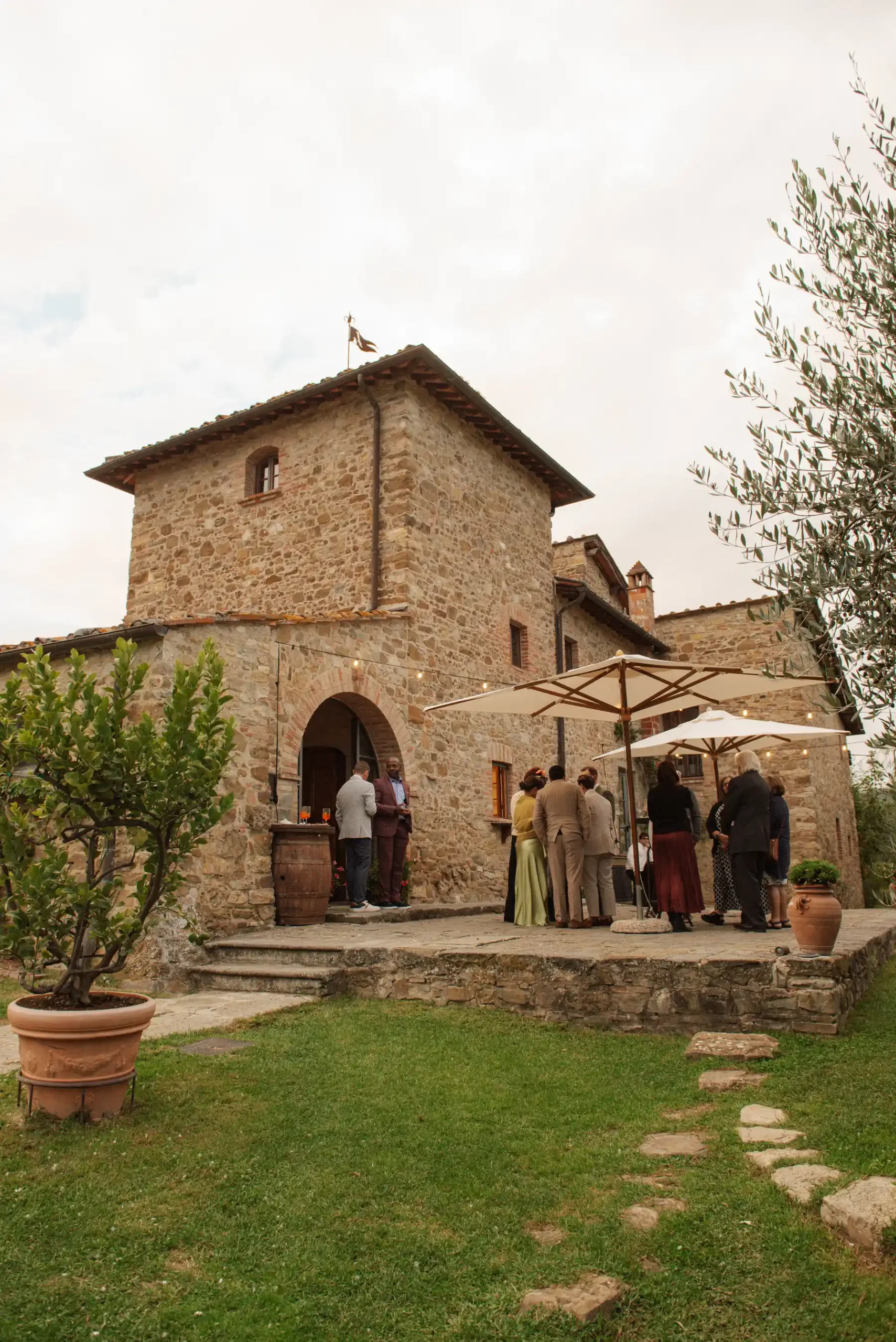 Guests mingle on the stone terrace of Casa Cornacchi in Arezzo, Tuscany, under cream market umbrellas at dusk.