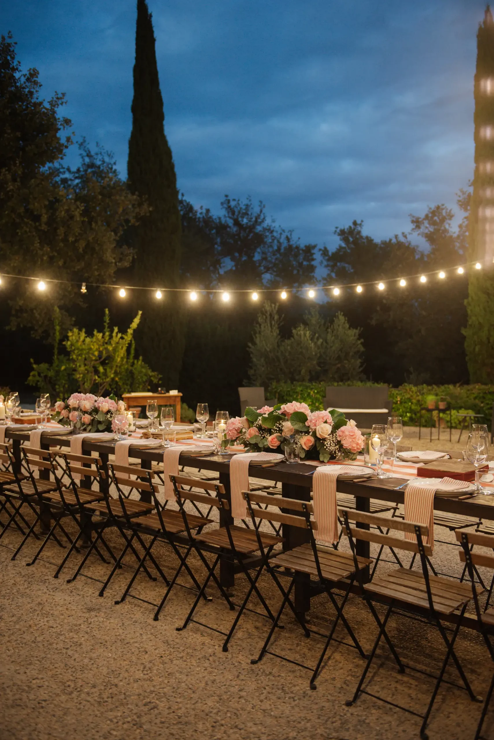 Outdoor reception tablescape at Casa Cornacchi in Tuscany with pink floral centerpieces, striped napkins and string lights at dusk.
