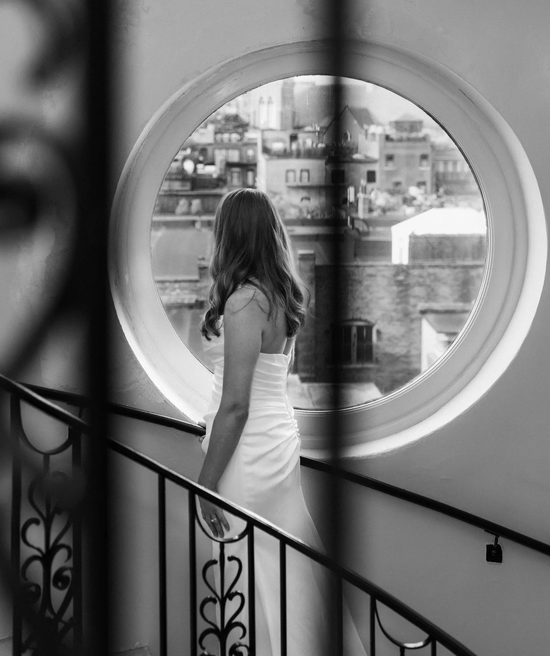 Bride Stephanie Tillman gazes through a round porthole window at Hampshire House in Boston, wearing a strapless white gown.