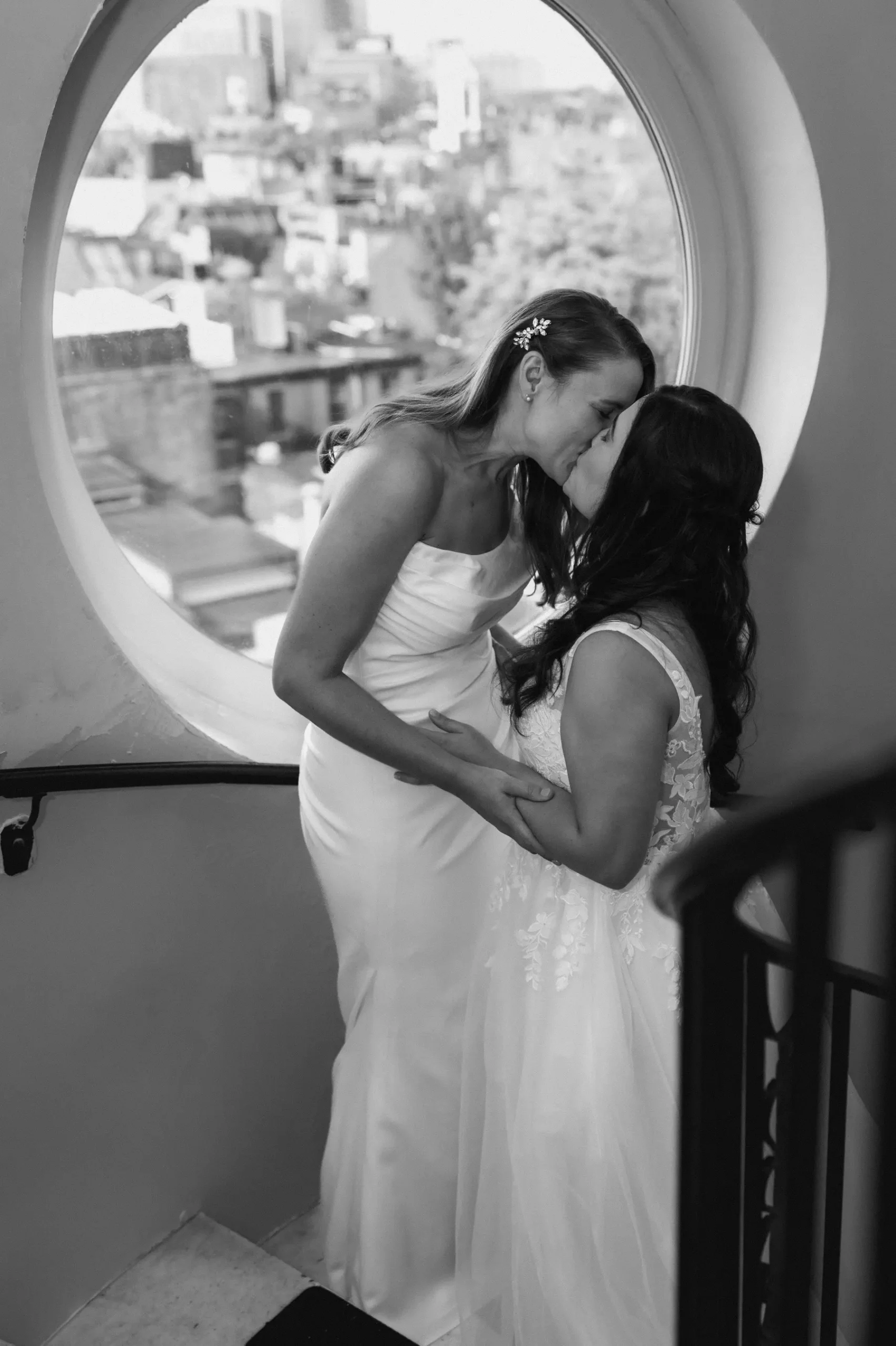 Brides Ashton Hattori and Stephanie Tillman share a kiss in wedding gowns by a round window at Hampshire House, Boston.