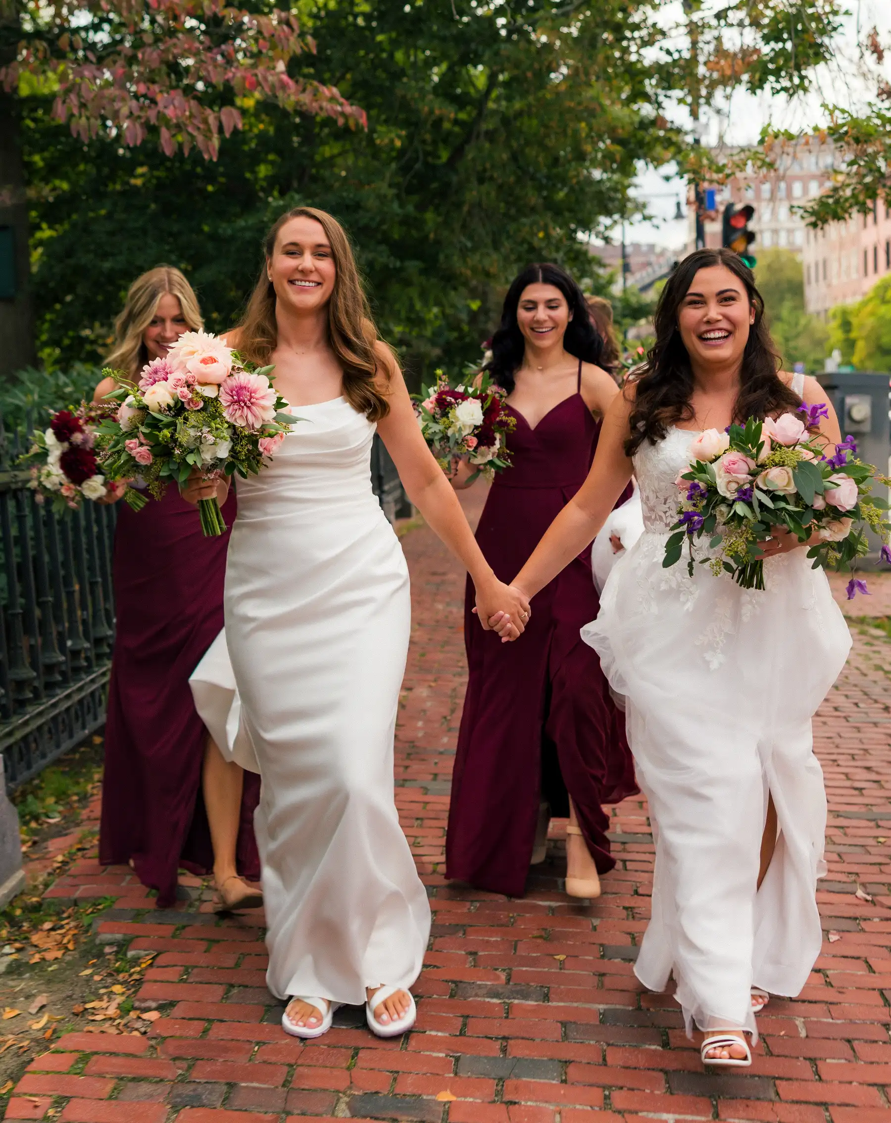 Brides Ashton Hattori and Stephanie Tillman hold hands and walk with bridesmaids on a brick path in Boston.