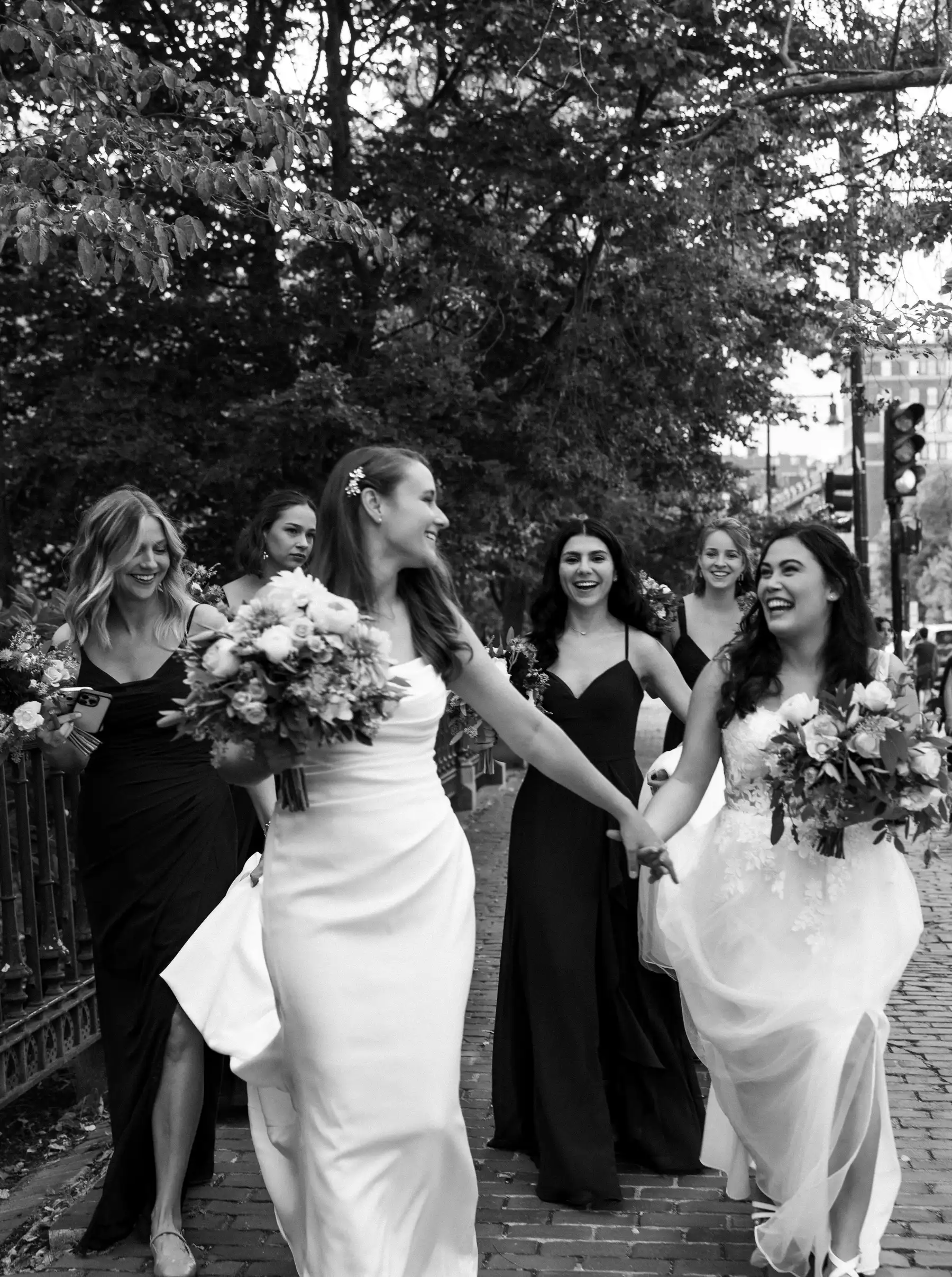 Brides Ashton Hattori and Stephanie Tillman hold hands and laugh with bridesmaids on cobblestone steps in Boston.