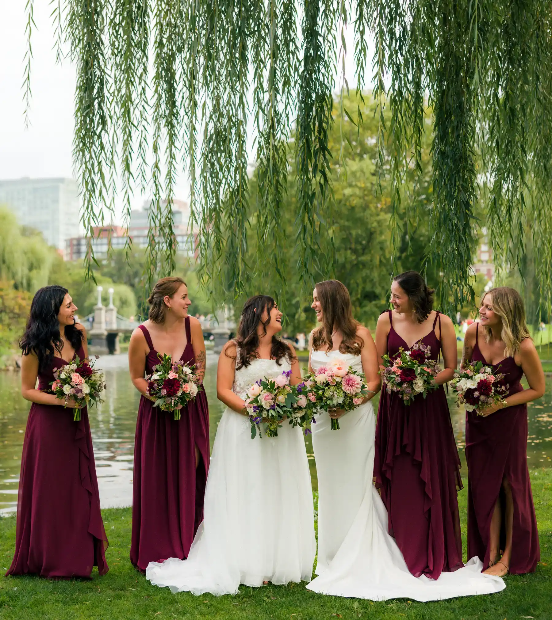 Brides Ashton Hattori and Stephanie Tillman laugh with four bridesmaids in burgundy gowns beneath a weeping willow in Boston.