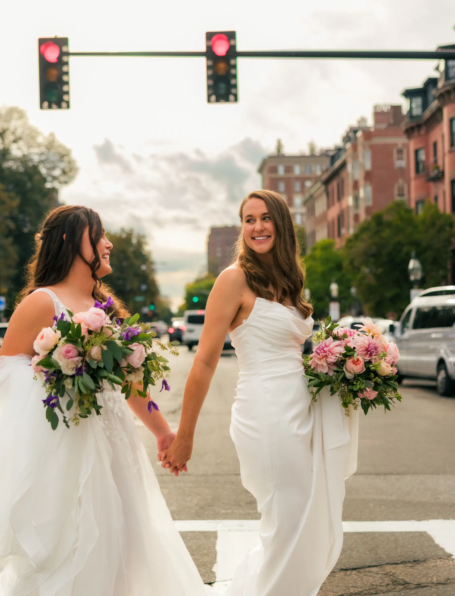 Brides Ashton Hattori and Stephanie Tillman hold hands crossing a Boston street in white wedding gowns with pink bouquets.