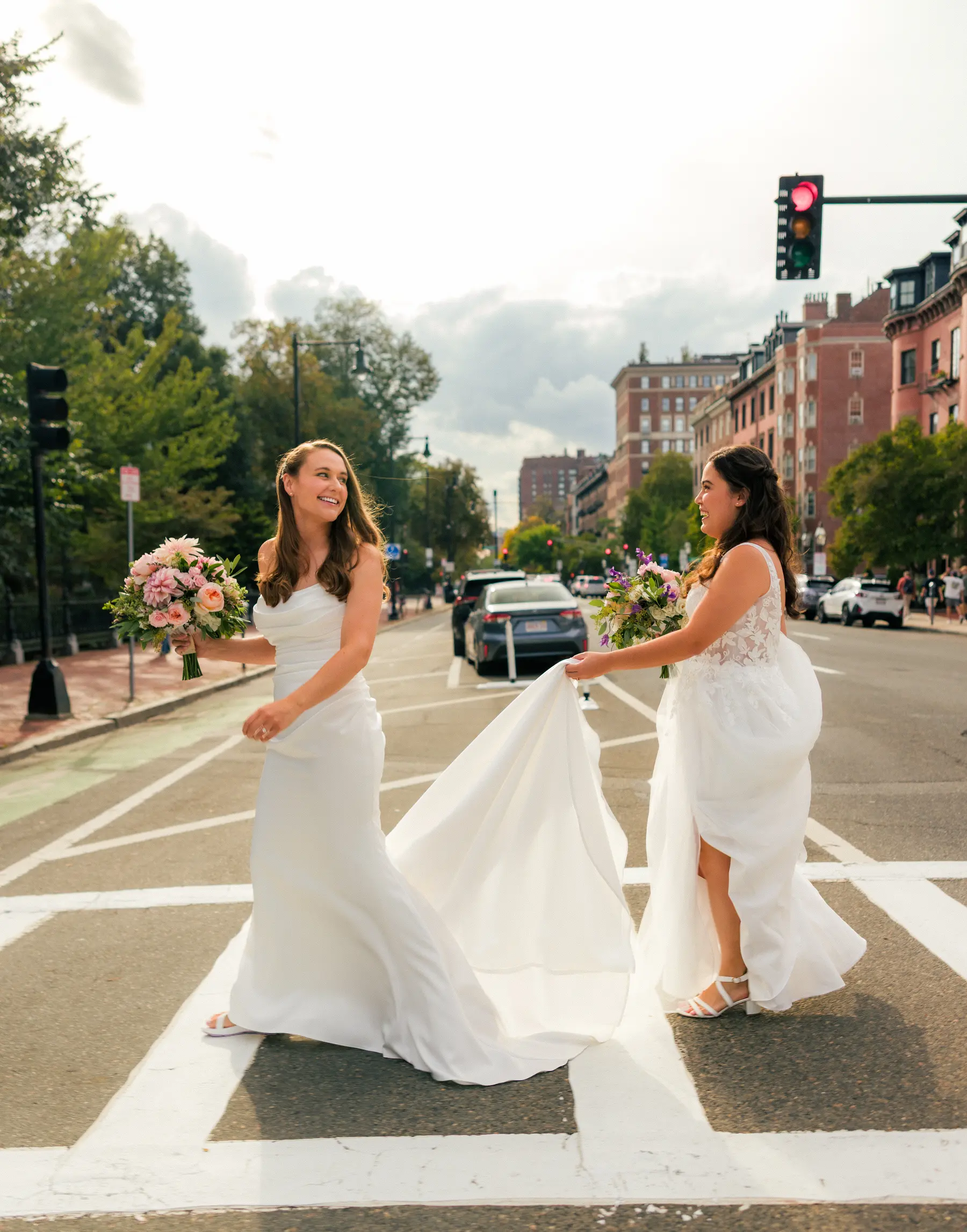 Brides Ashton Hattori and Stephanie Tillman cross a sunlit Boston street in white gowns, bouquets in hand.