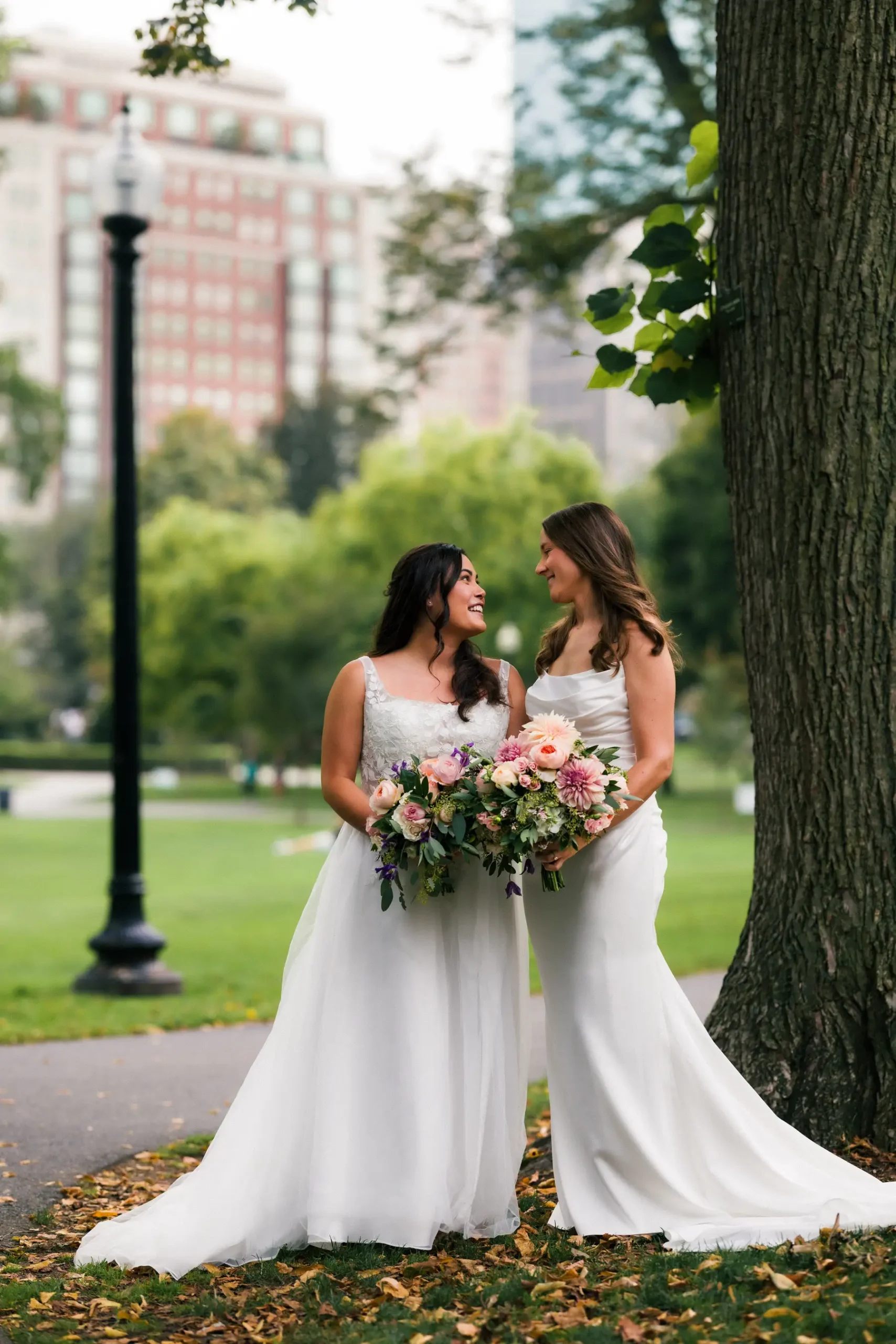 Brides Ashton Hattori and Stephanie Tillman smile at each other in white gowns holding bouquets in a Boston park.