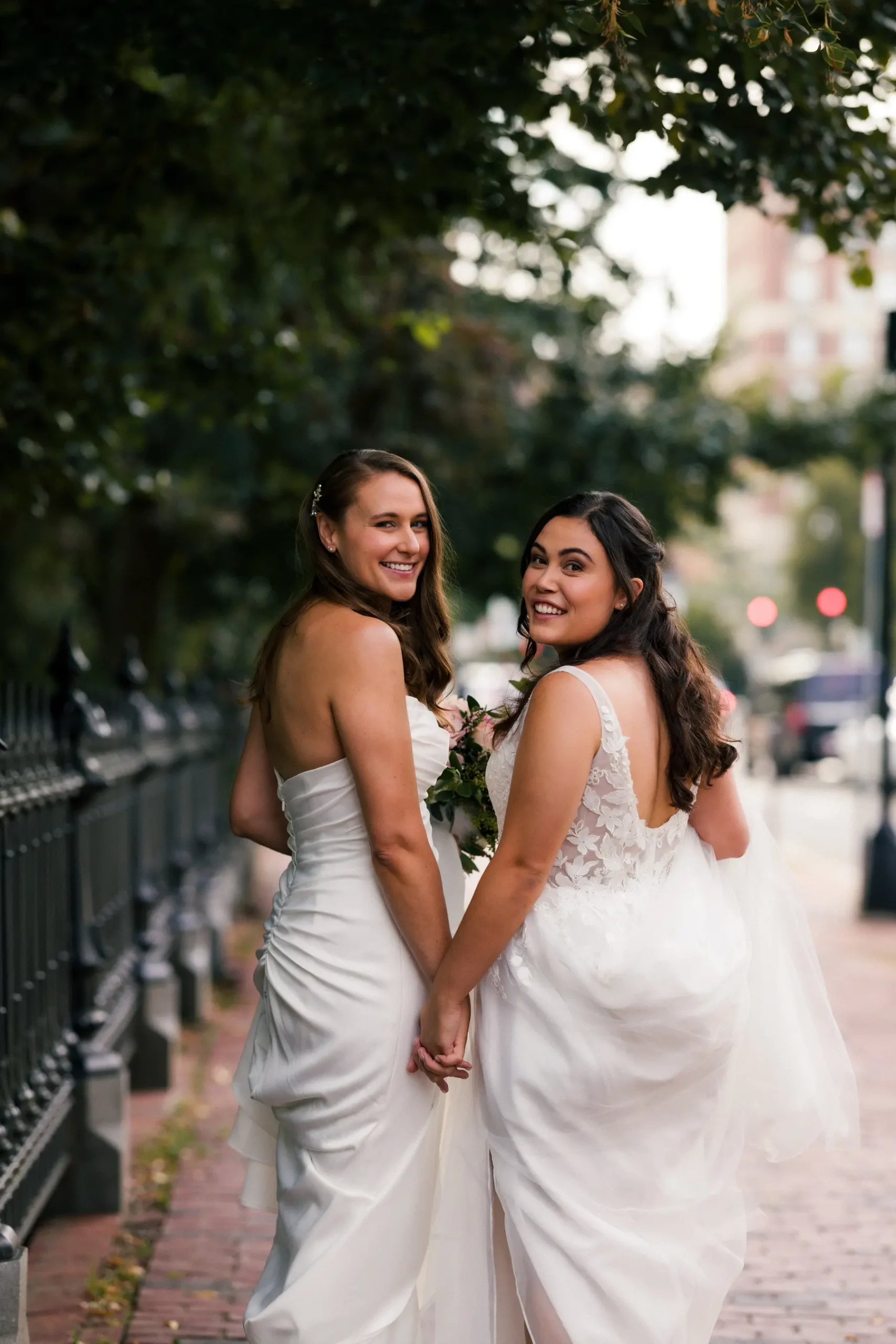 Brides Ashton Hattori and Stephanie Tillman hold hands in white gowns on a tree-lined Boston sidewalk near Hampshire House.