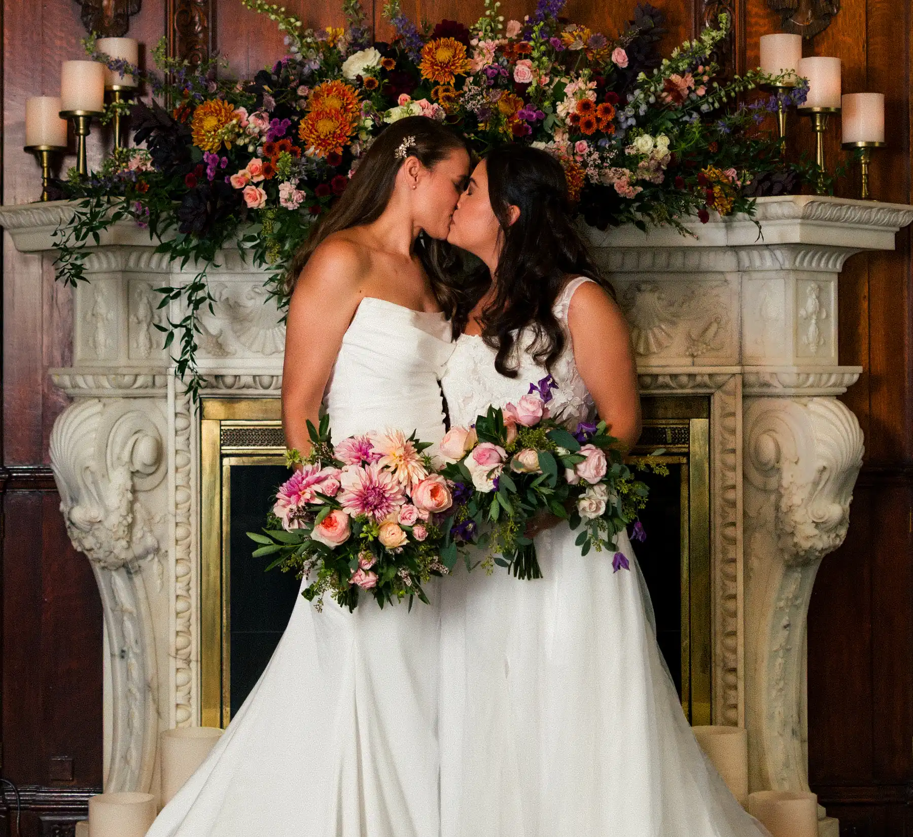 Brides Ashton Hattori and Stephanie Tillman share a kiss in white gowns before a floral-adorned marble fireplace at Hampshire House in Boston.