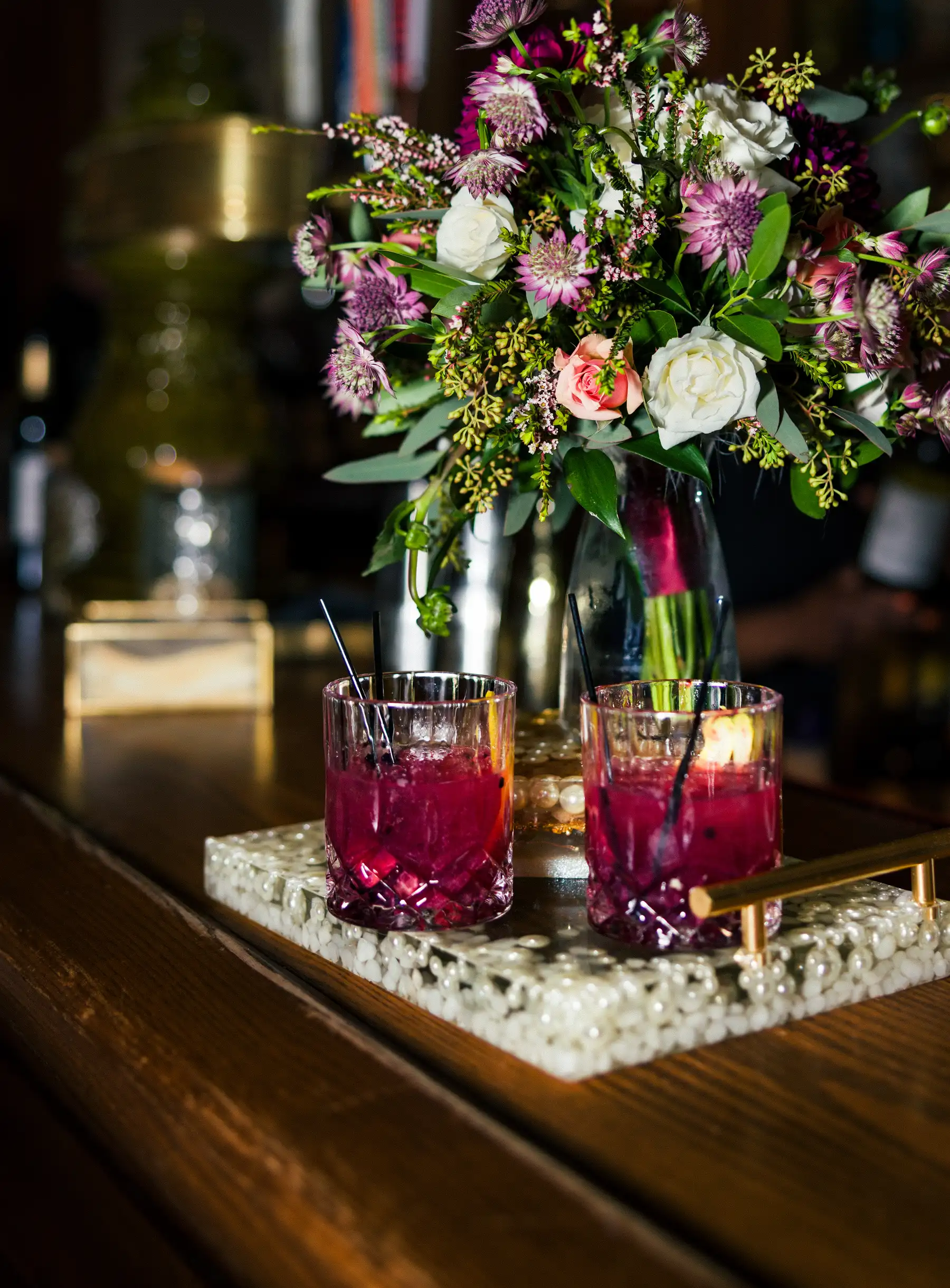 Two deep crimson cocktails on a pearl tray beside a lush floral arrangement at Hampshire House in Boston.