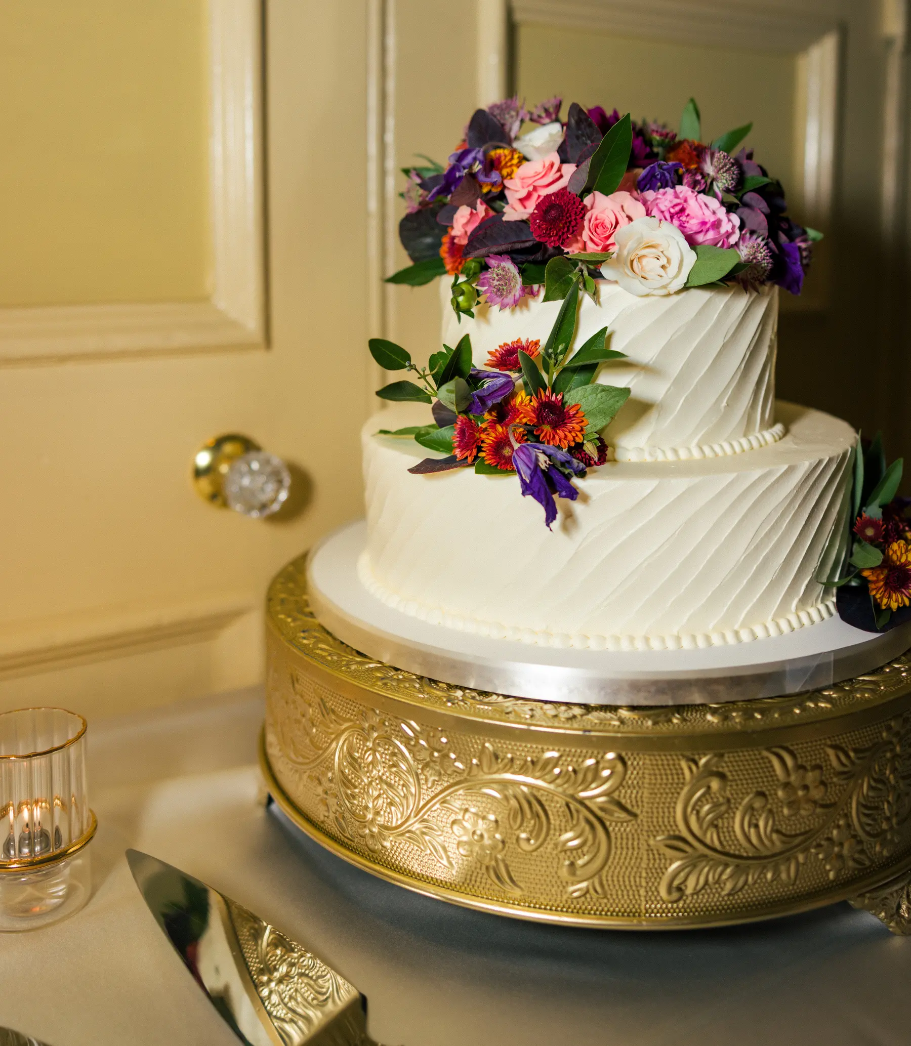 A three-tier white wedding cake adorned with colorful flowers on a gold stand at Hampshire House in Boston.