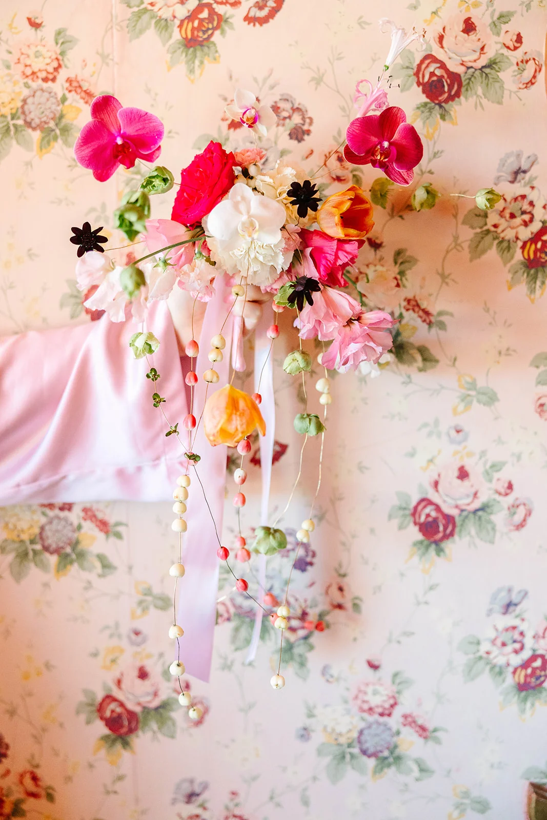 A lush cascading bridal bouquet with pink orchids, tulips and trailing berry stems against floral wallpaper at Madonna Inn.