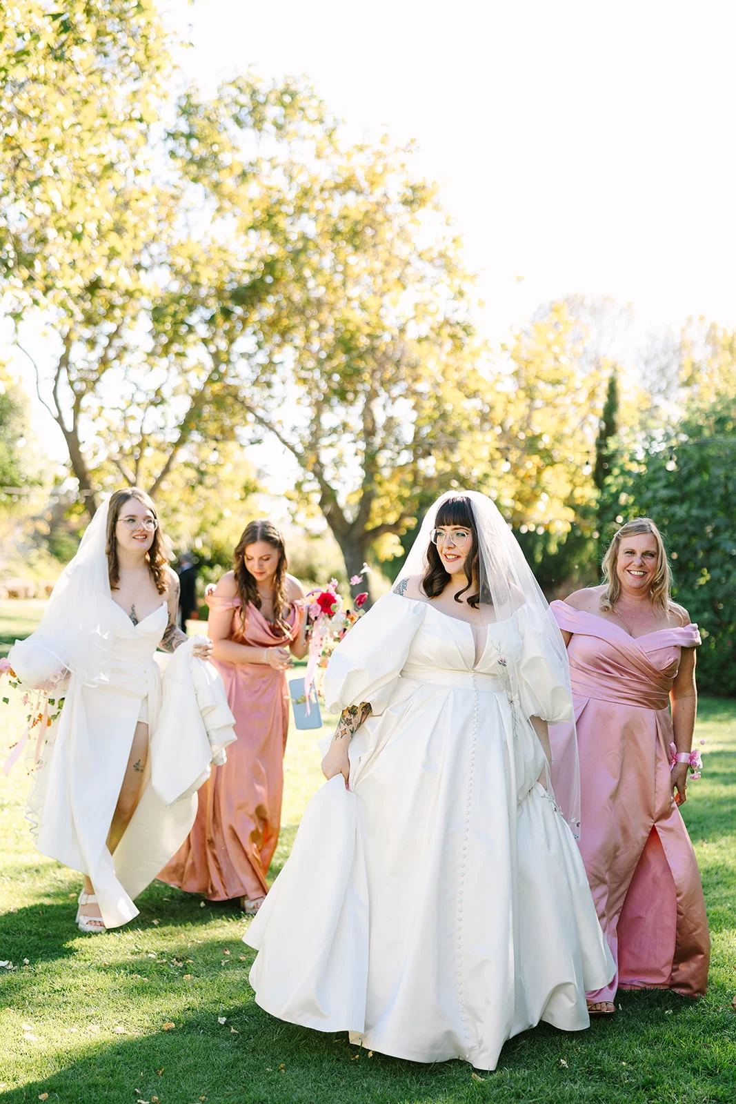 Bride Mimi walks with bridesmaids in pink gowns across a sunny lawn at Madonna Inn in San Luis Obispo, California.