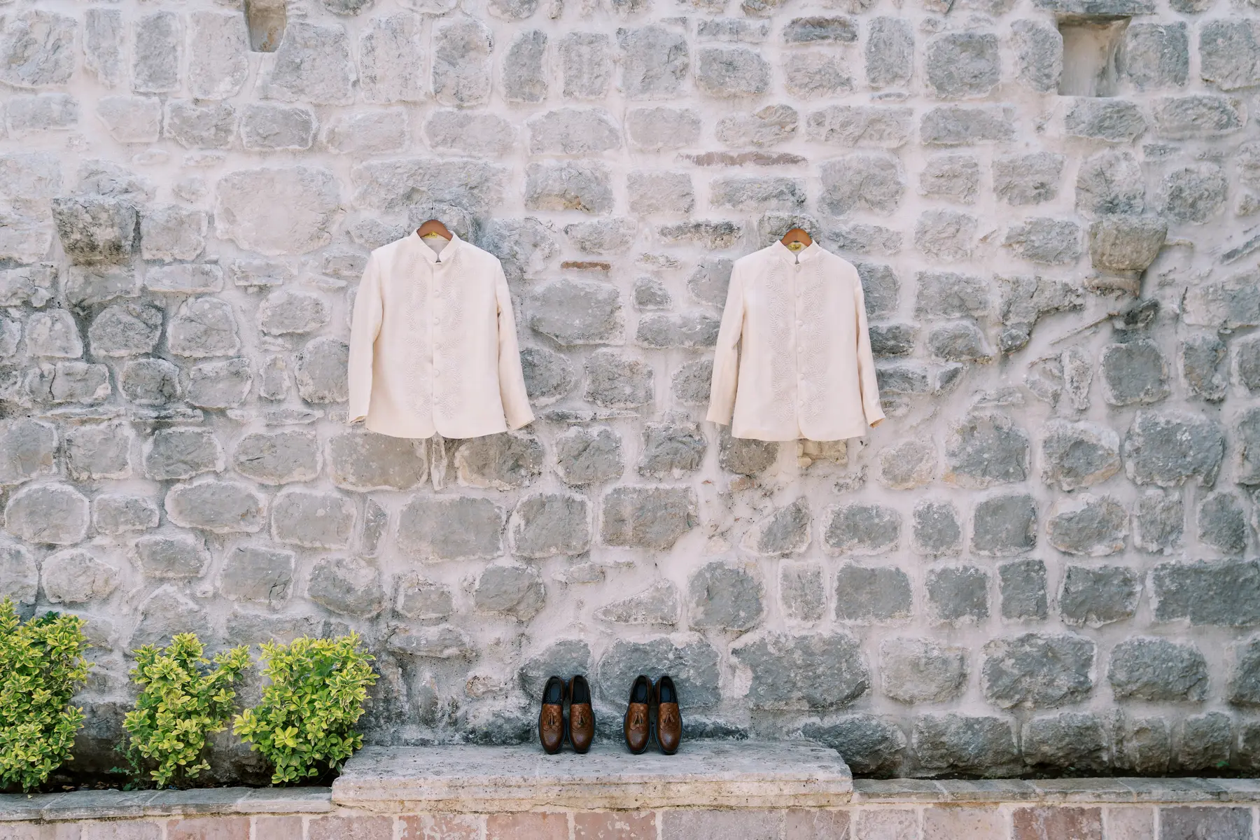 Two ivory Mandarin-collar suits on hangers and brown tassel loafers displayed against a stone wall at Monte Bay Retreat in Perast, Montenegro.