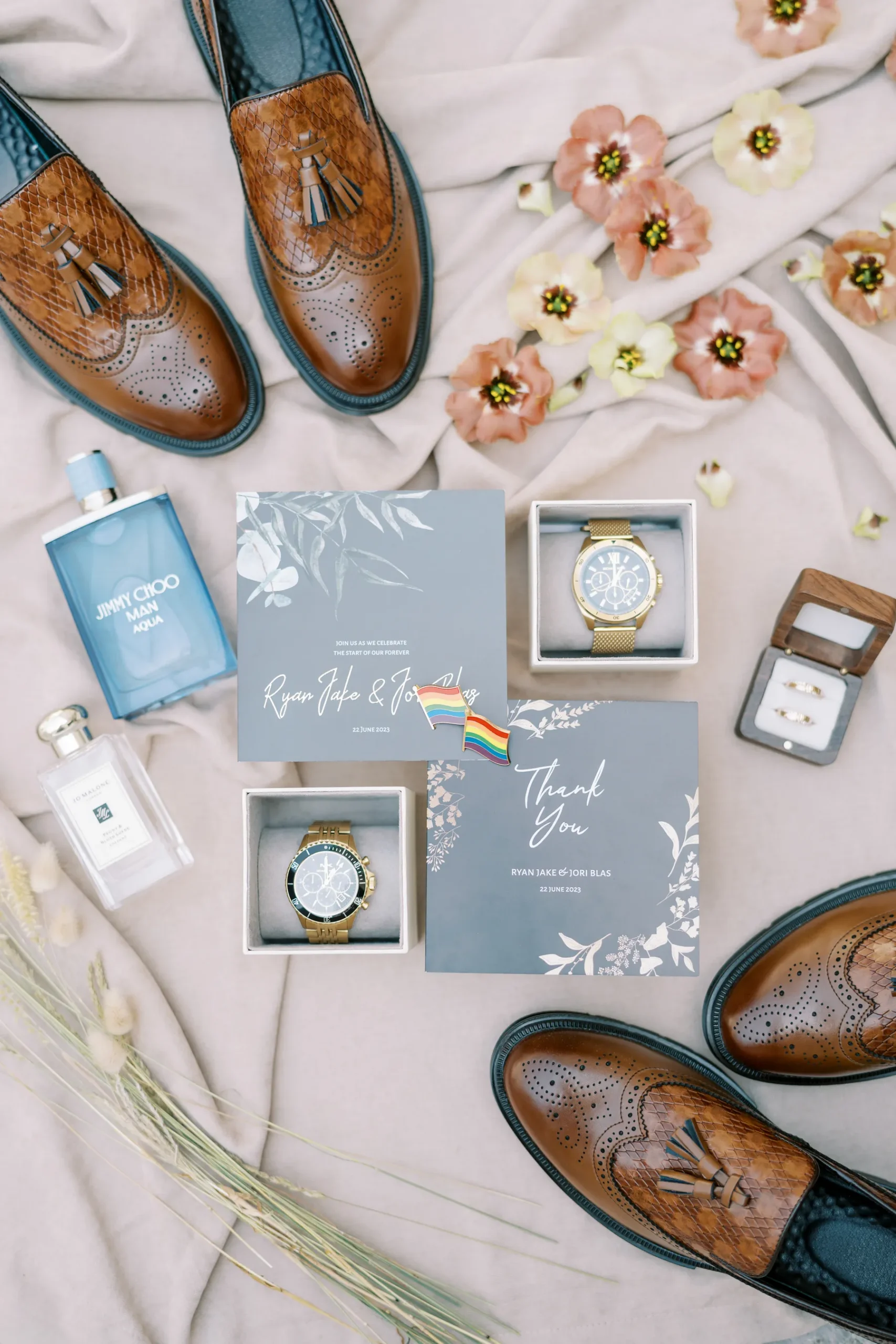 Wedding flat lay for Ryan Jake and Jori Blas featuring blue stationery, gold watches, brown loafers, and a rainbow pride pin.
