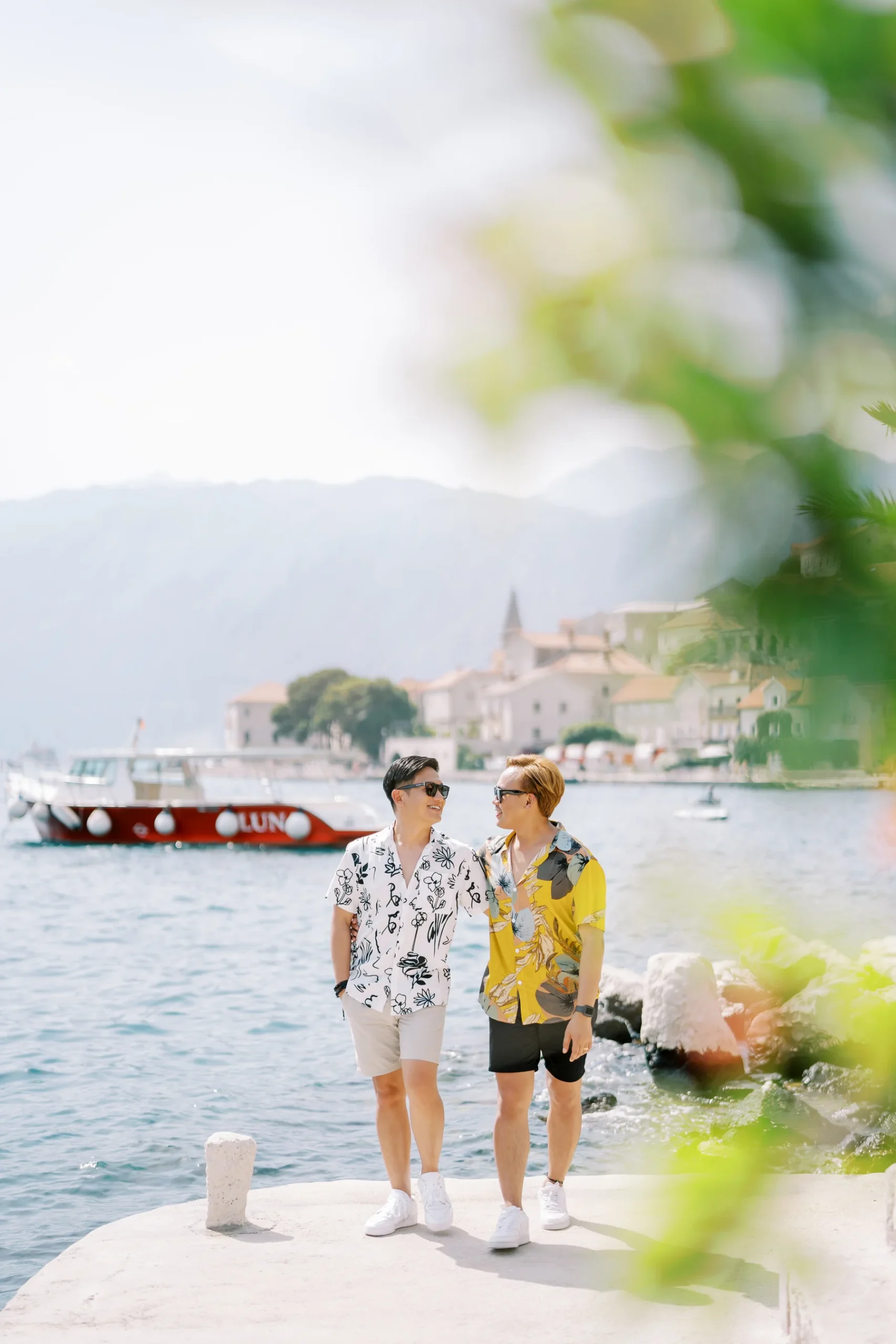 Ryan Jake and Jori smile at each other on a sunlit pier in Perast, Montenegro, wearing colorful floral shirts.