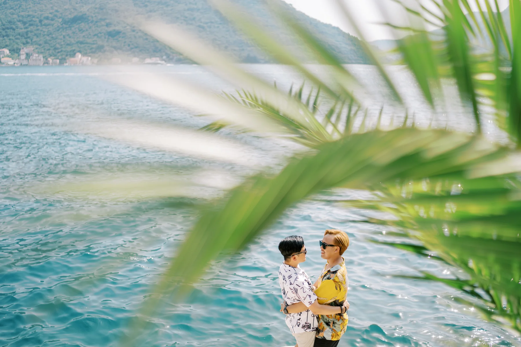 Ryan Jake and Jori embrace by the turquoise waters of Monte Bay Retreat in Perast, Montenegro, framed by palm fronds.