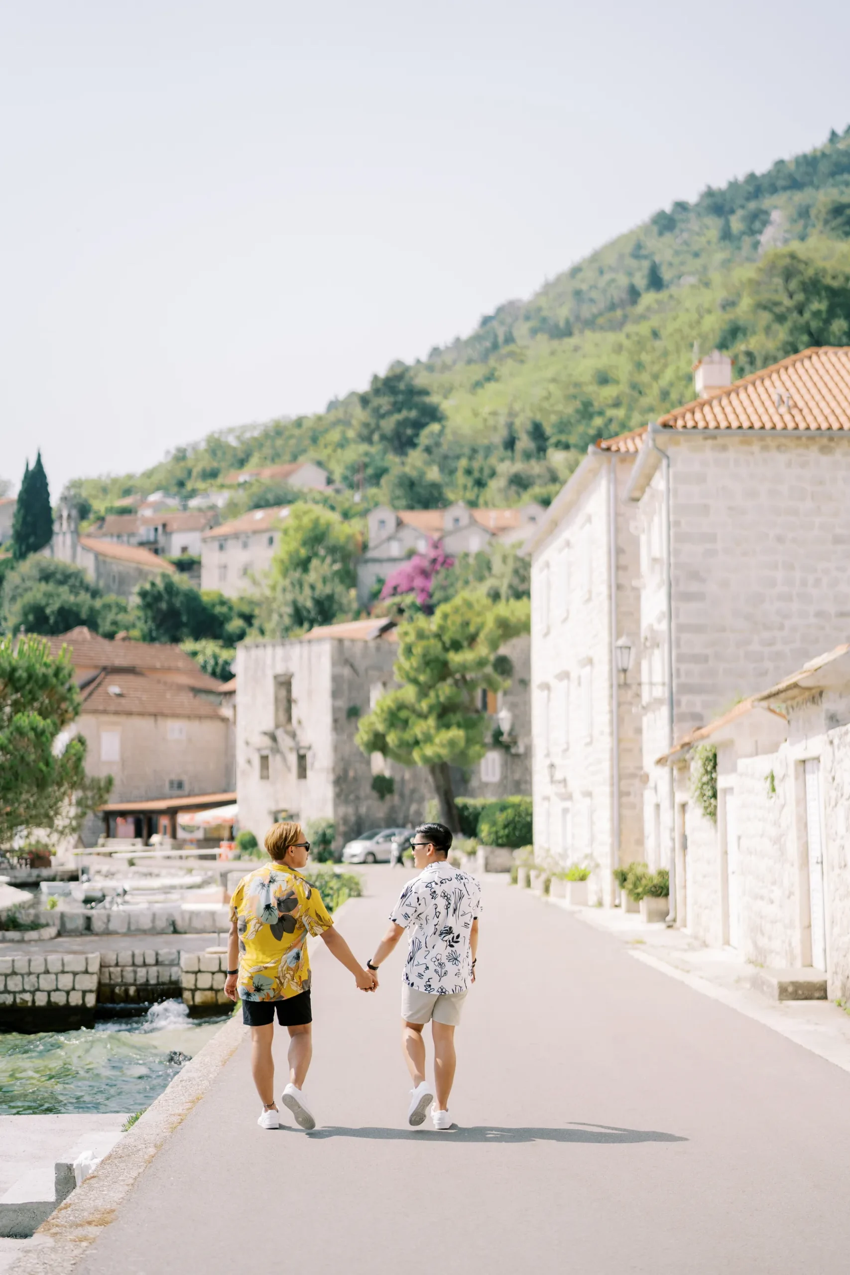 Ryan Jake and Jori hold hands walking along a sunlit stone promenade in Perast, Montenegro.
