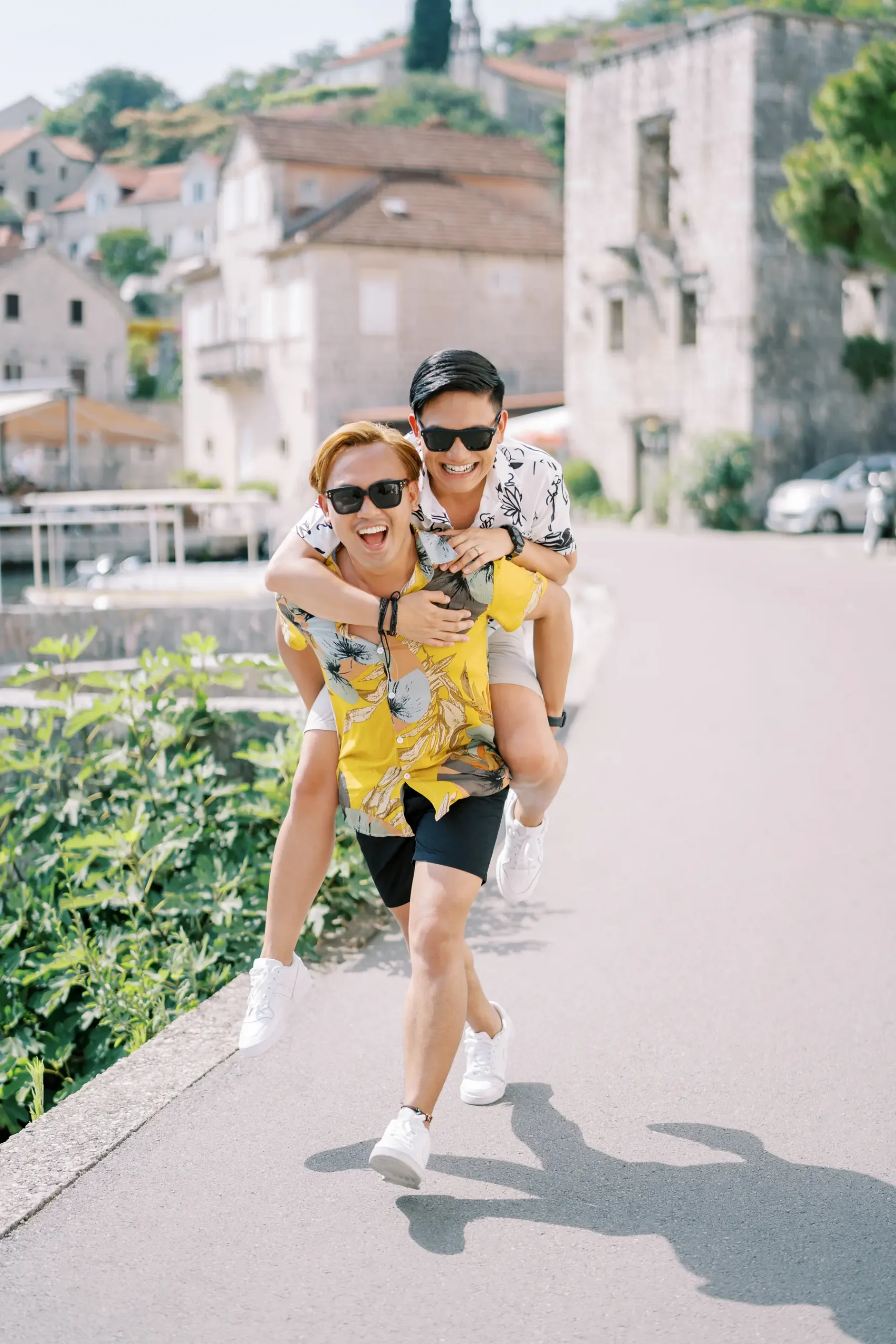 Ryan and Jori laugh during a piggyback ride along a sunlit promenade in Perast, Montenegro.