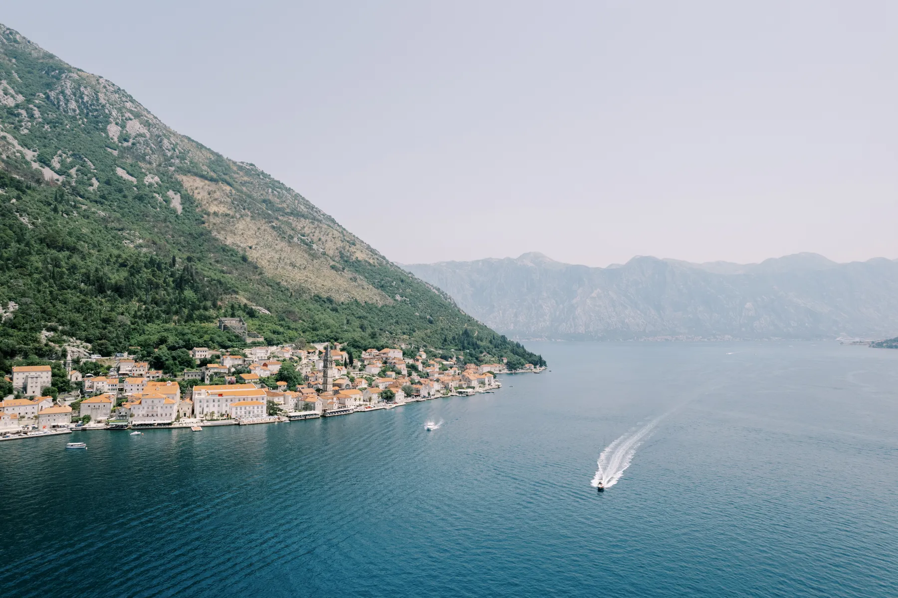 Aerial view of Perast, Montenegro's Bay of Kotor shoreline with terracotta-roofed buildings and a speedboat wake.