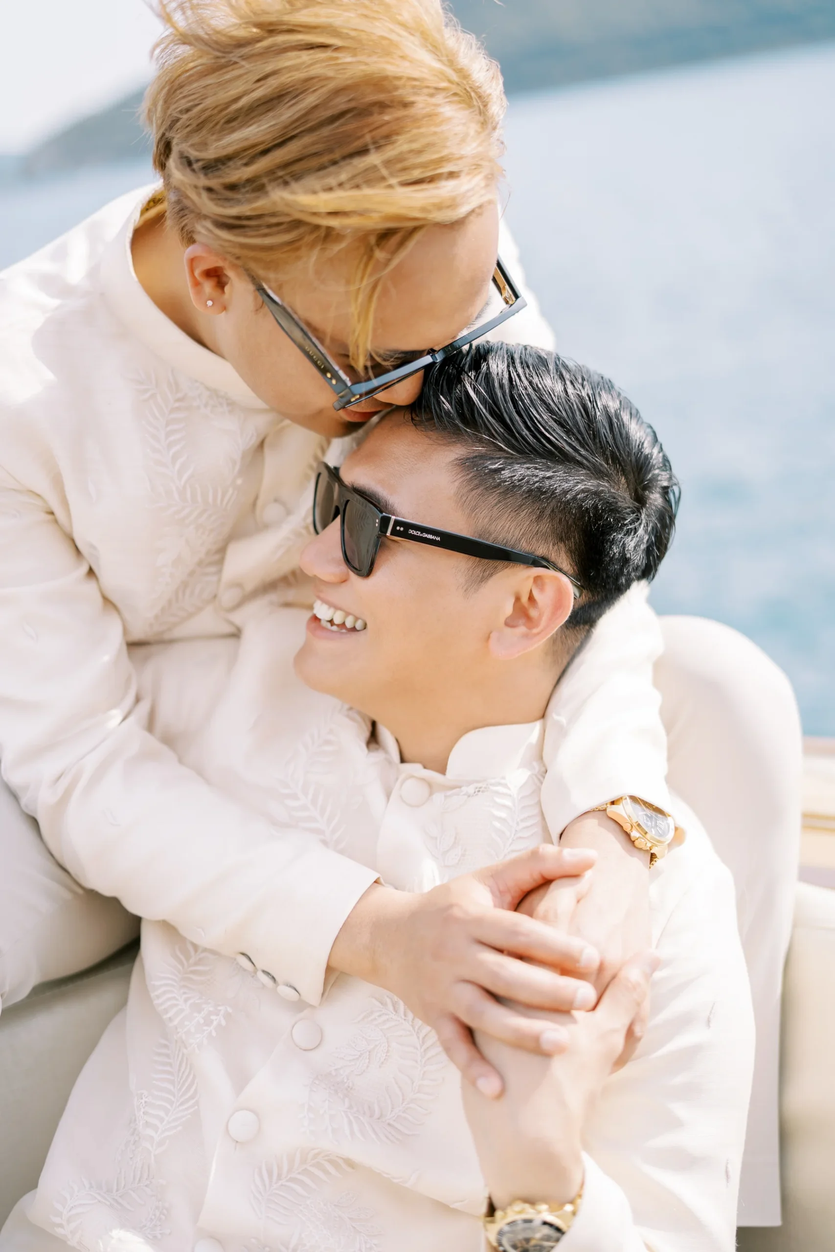 Ryan Jake and Jori embrace on a boat near Monte Bay Retreat in Perast, Montenegro, both wearing matching cream embroidered suits.