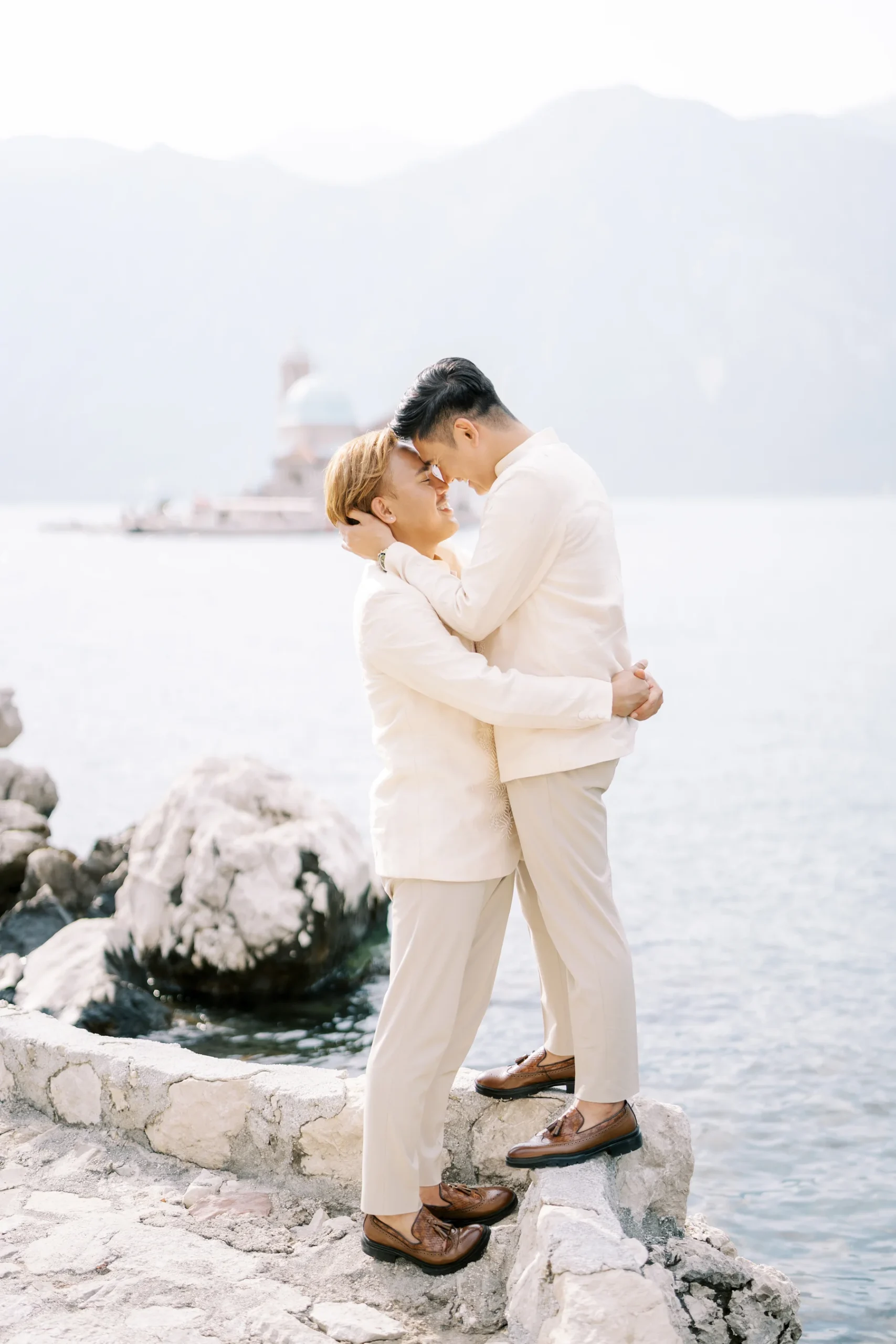 Ryan Jake and Jori embrace in cream suits on rocky shore at Monte Bay Retreat in Perast, Montenegro, foreheads touching.